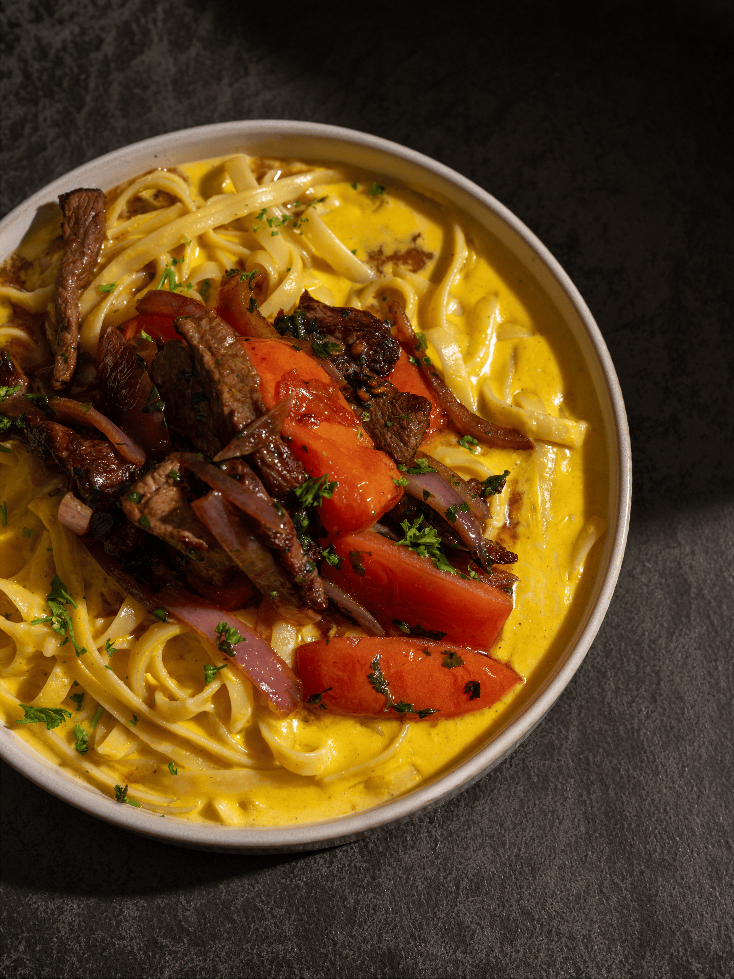 A bowl of fettuccine pasta in a creamy huancaina sauce, topped with grilled beef, sliced tomatoes, and herbs, on a dark surface. Lomo saltado