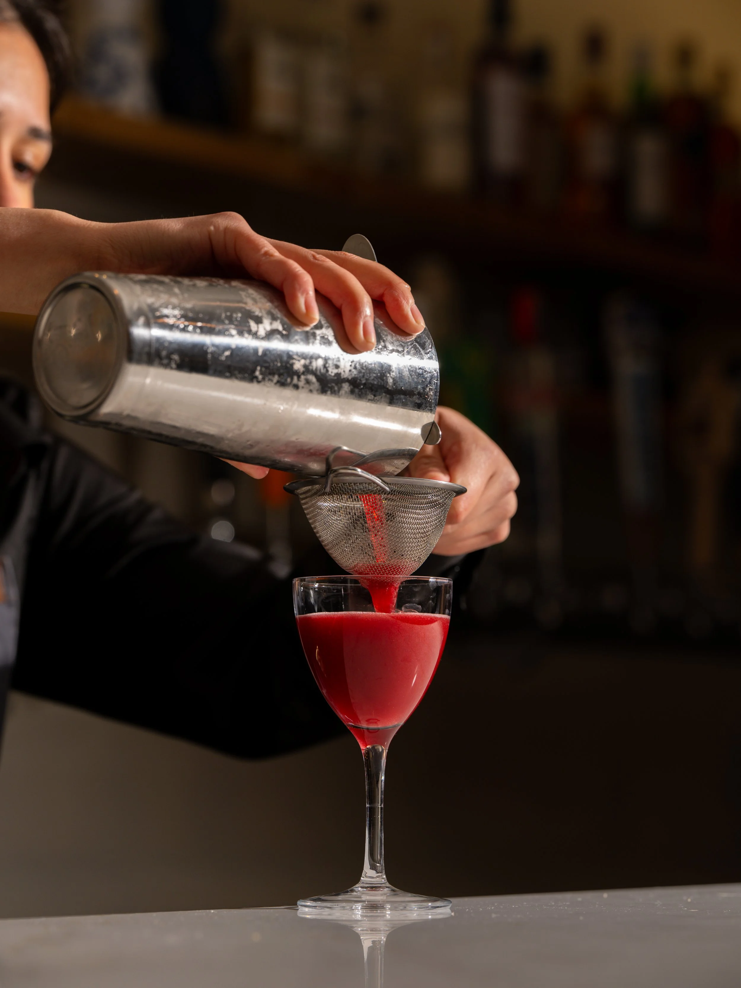 A person pouring a red cocktail through a fine mesh strainer into a stemmed glass. Pachas elixir cocktail at Mercado Restaurant