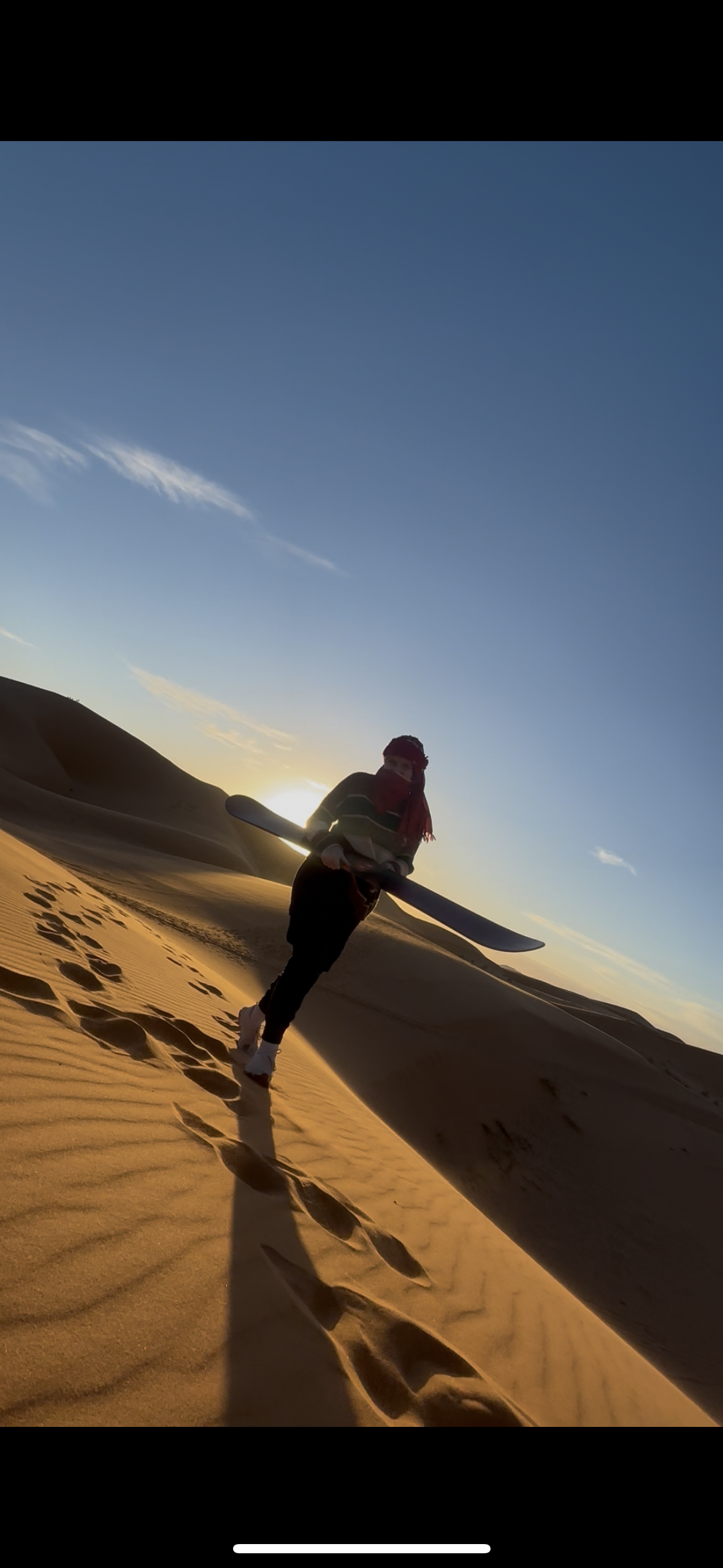 Person walking in desert at sunset, holding a kayak paddle, with footprints and sand dunes in the background.