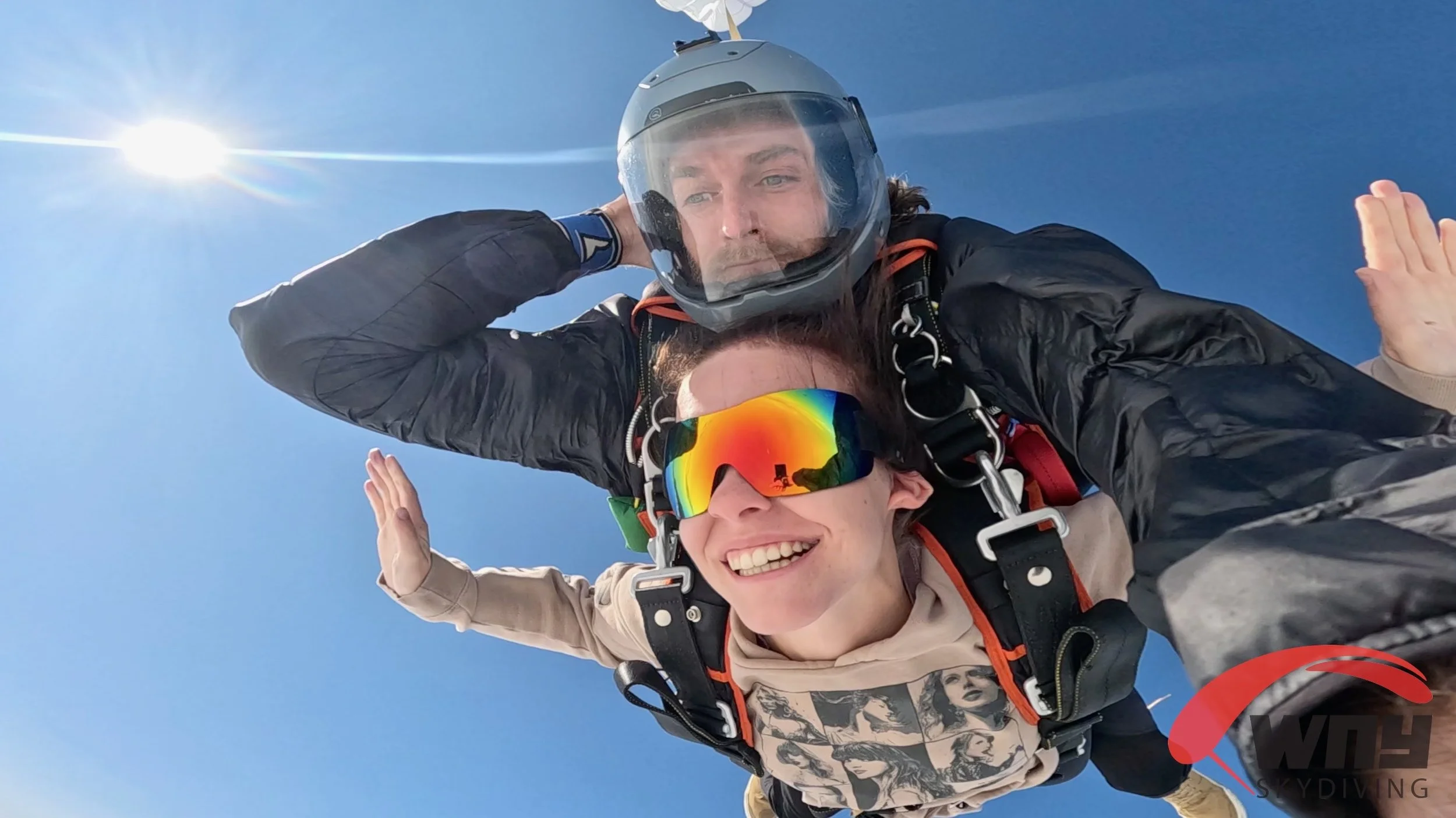 Two skydivers in freefall, one wearing a gray helmet and the other with rainbow-colored goggles, sky is clear blue with the sun shining brightly.