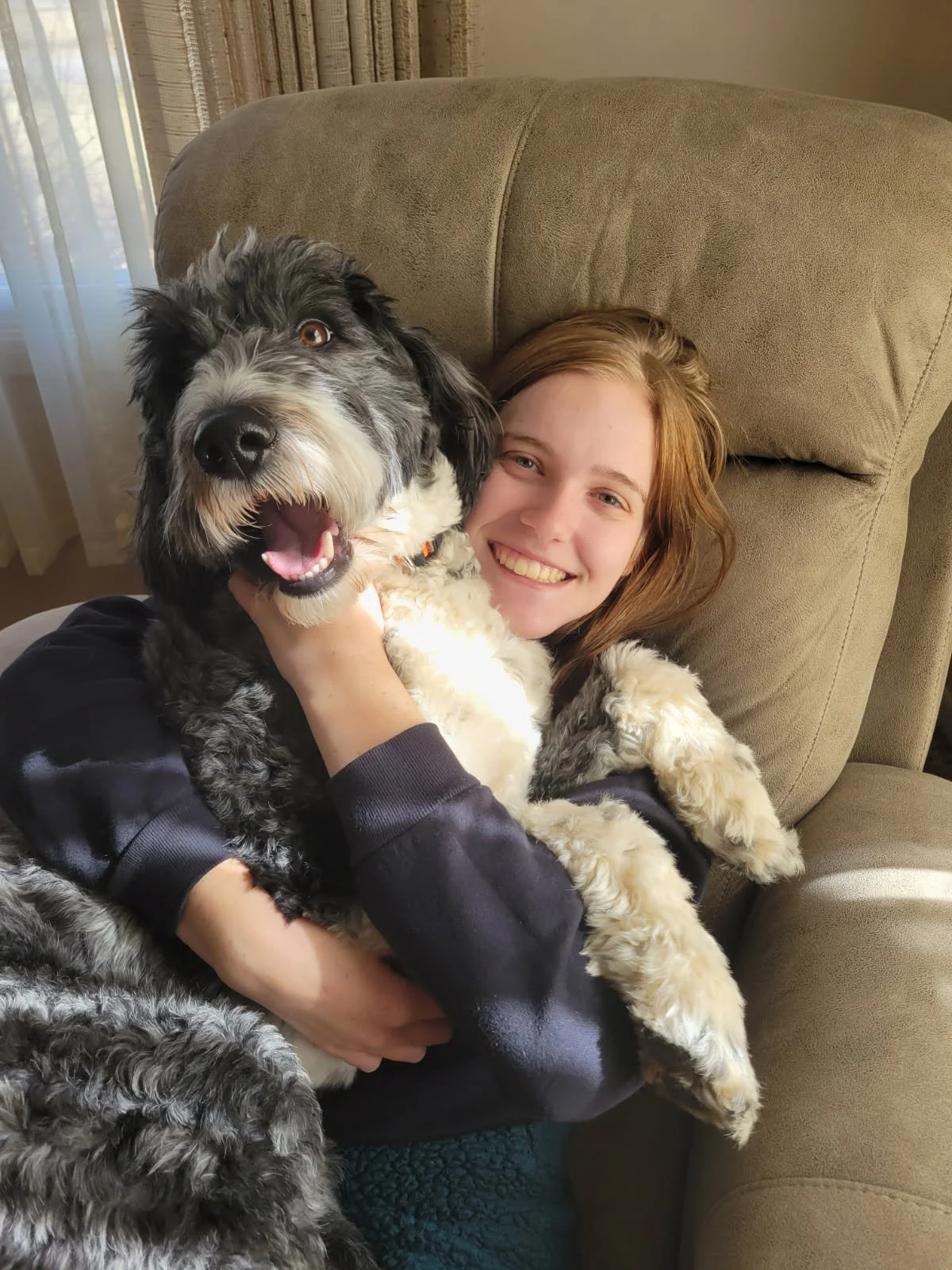 A young woman with brown hair smiling while holding a large, fluffy black and white dog in her arms, sitting on a beige sofa.