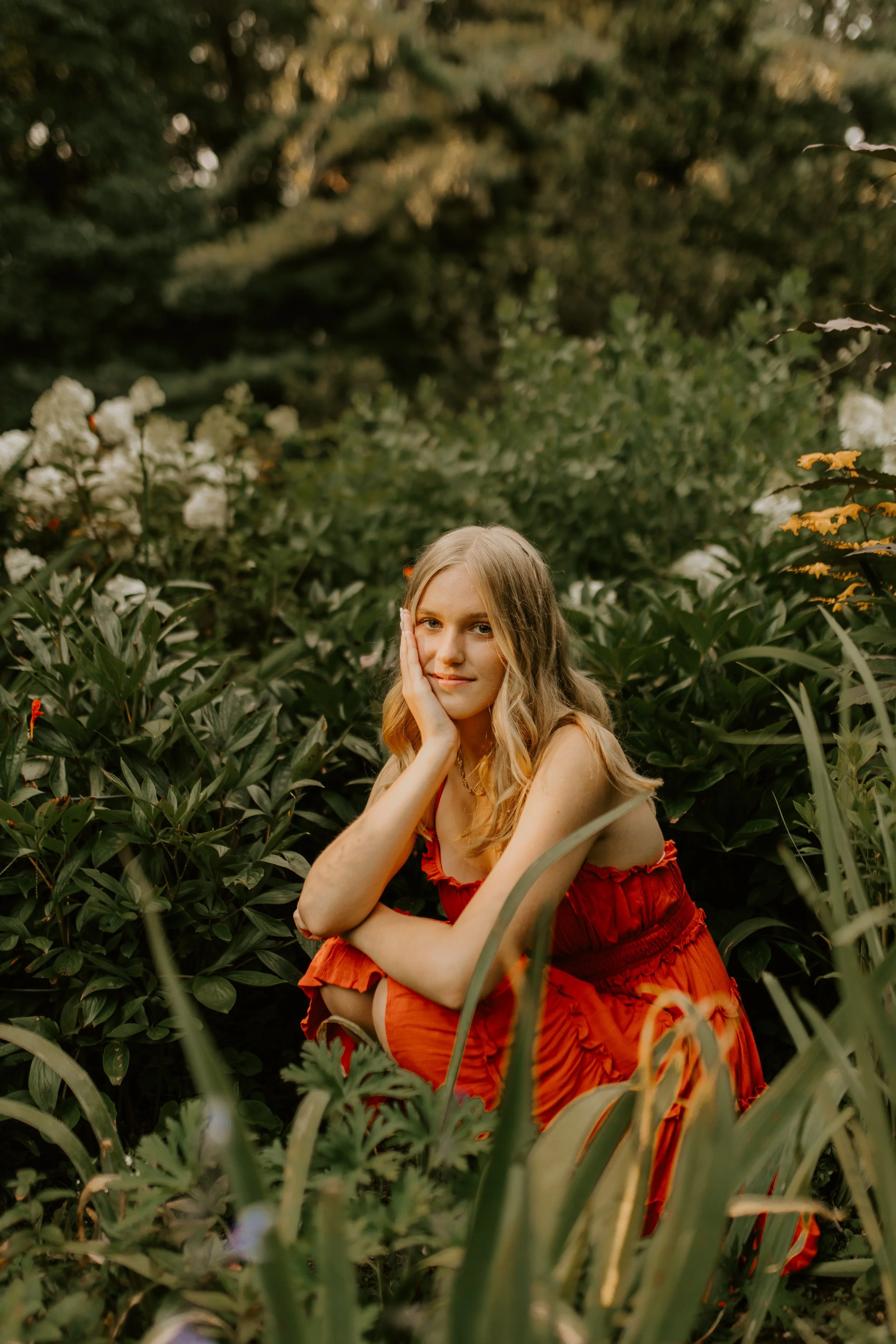 A young woman with long blonde hair, wearing a red dress, sitting among green plants and flowers in a natural outdoor setting.