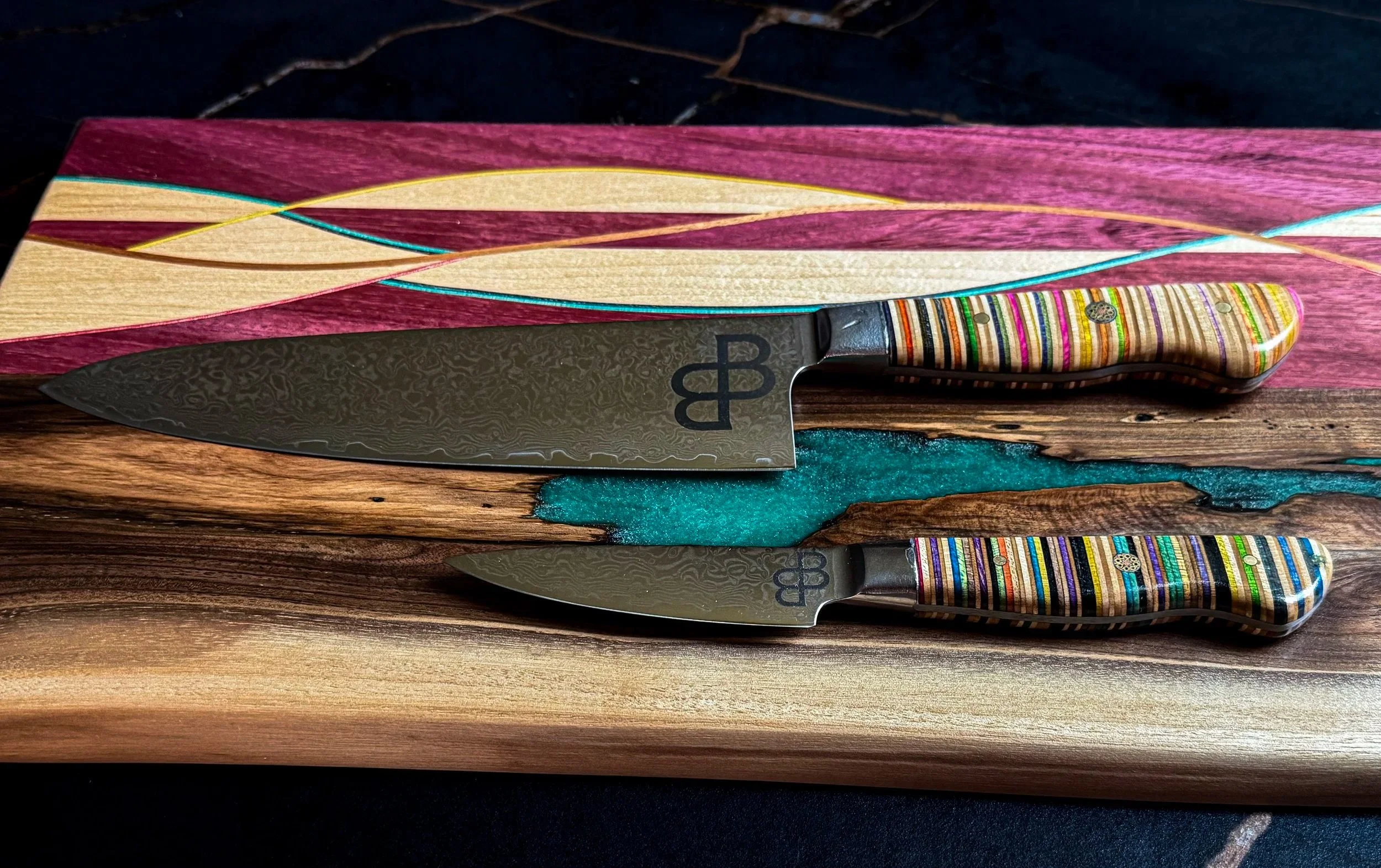 Handcrafted Damascus chef and paring knives with recycled skateboard handles displayed on artisan serving boards.