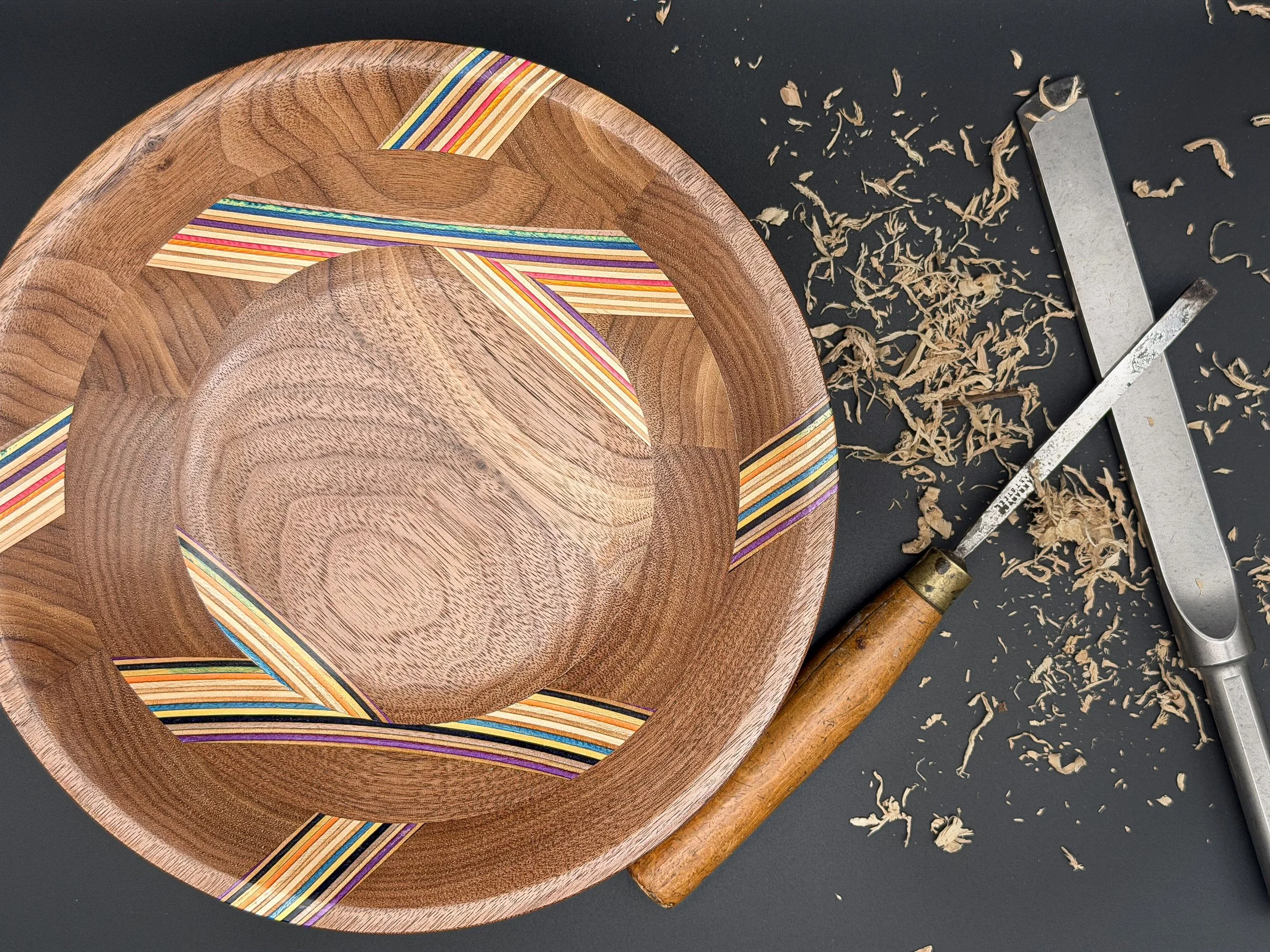 Close-up of a wooden bowl with colorful inlay designs, woodworking tools, and wood shavings on a black surface.