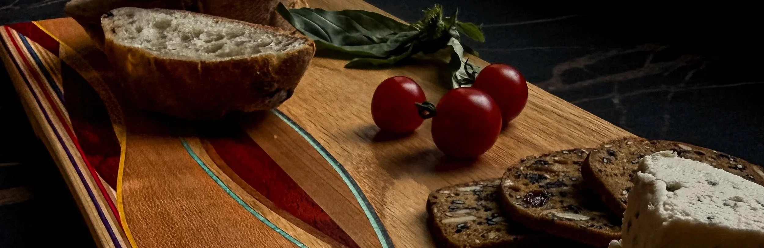 A wooden cutting board with three cherry tomatoes, some basil leaves, and slices of rustic bread topped with cheese. recycled skateboard woven inlay.