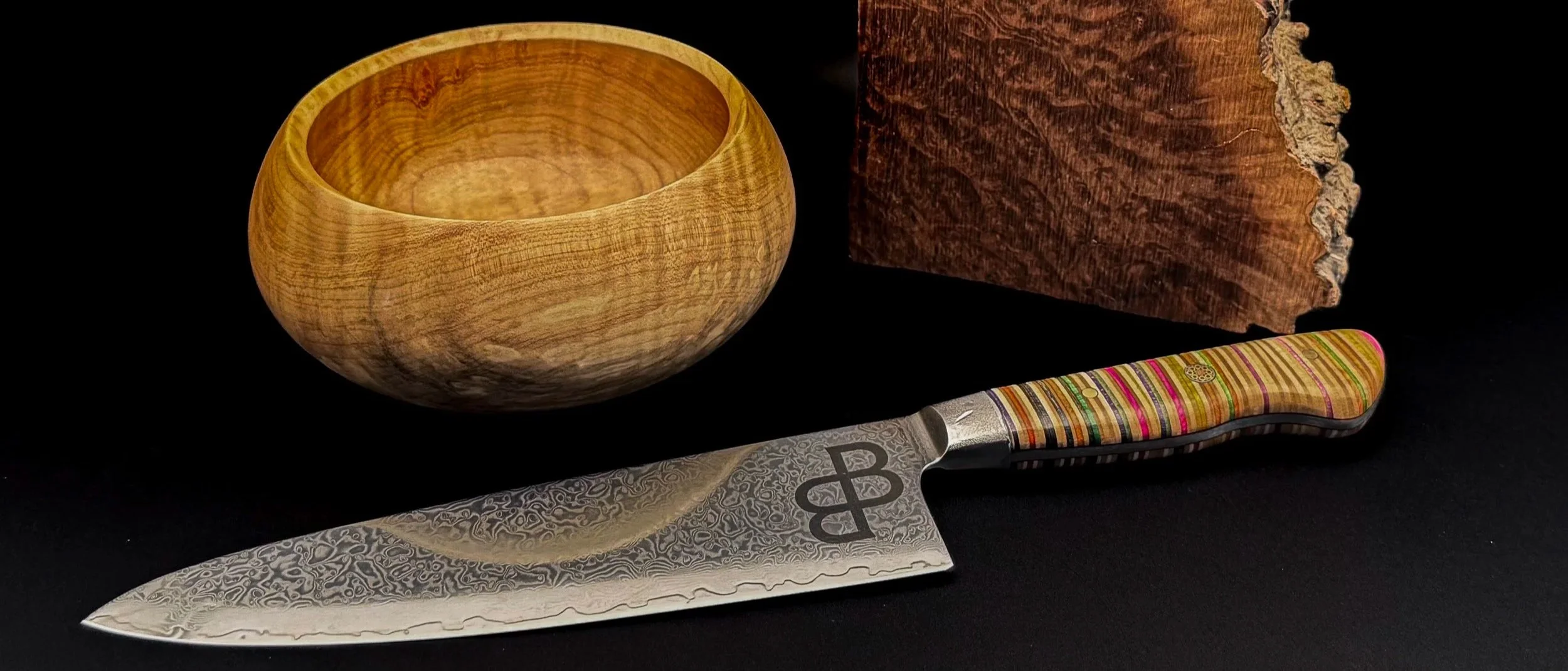 A wooden bowl, a piece of burl wood, a knife with colourful recycled skateboard handle, and the knife's blade with intricate Japanese Damascus steel patterns and a mark for blackburl works