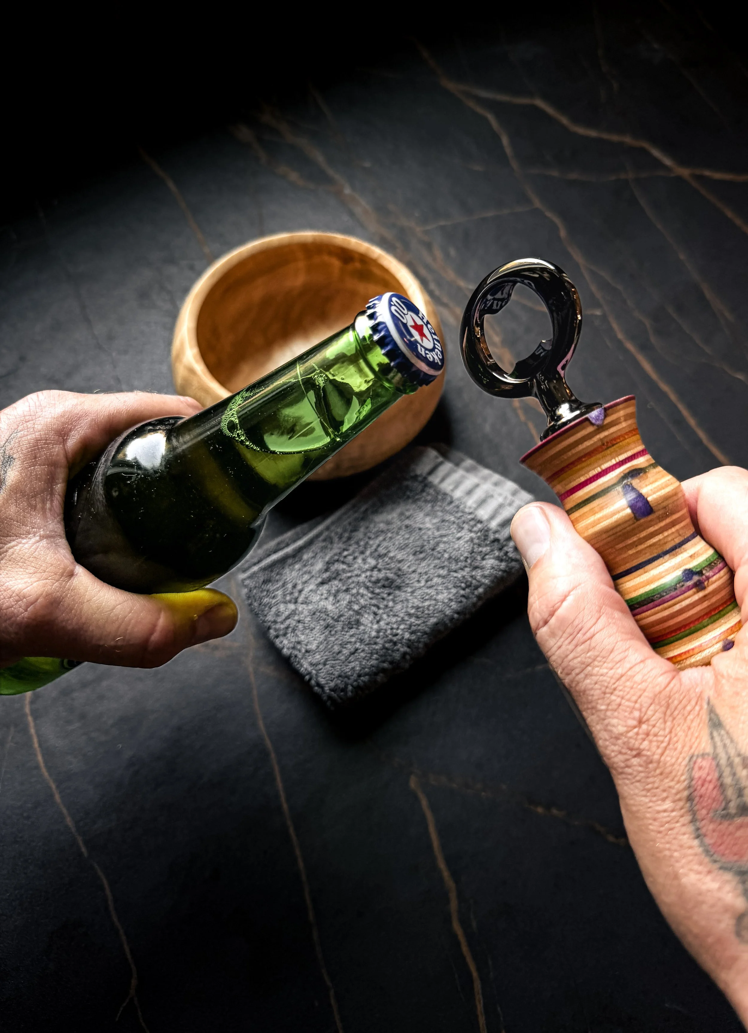 Person holding a green glass beer bottle and a wooden bottle opener  made from recycled skateboards over a dark surface with a wooden bowl nearby.