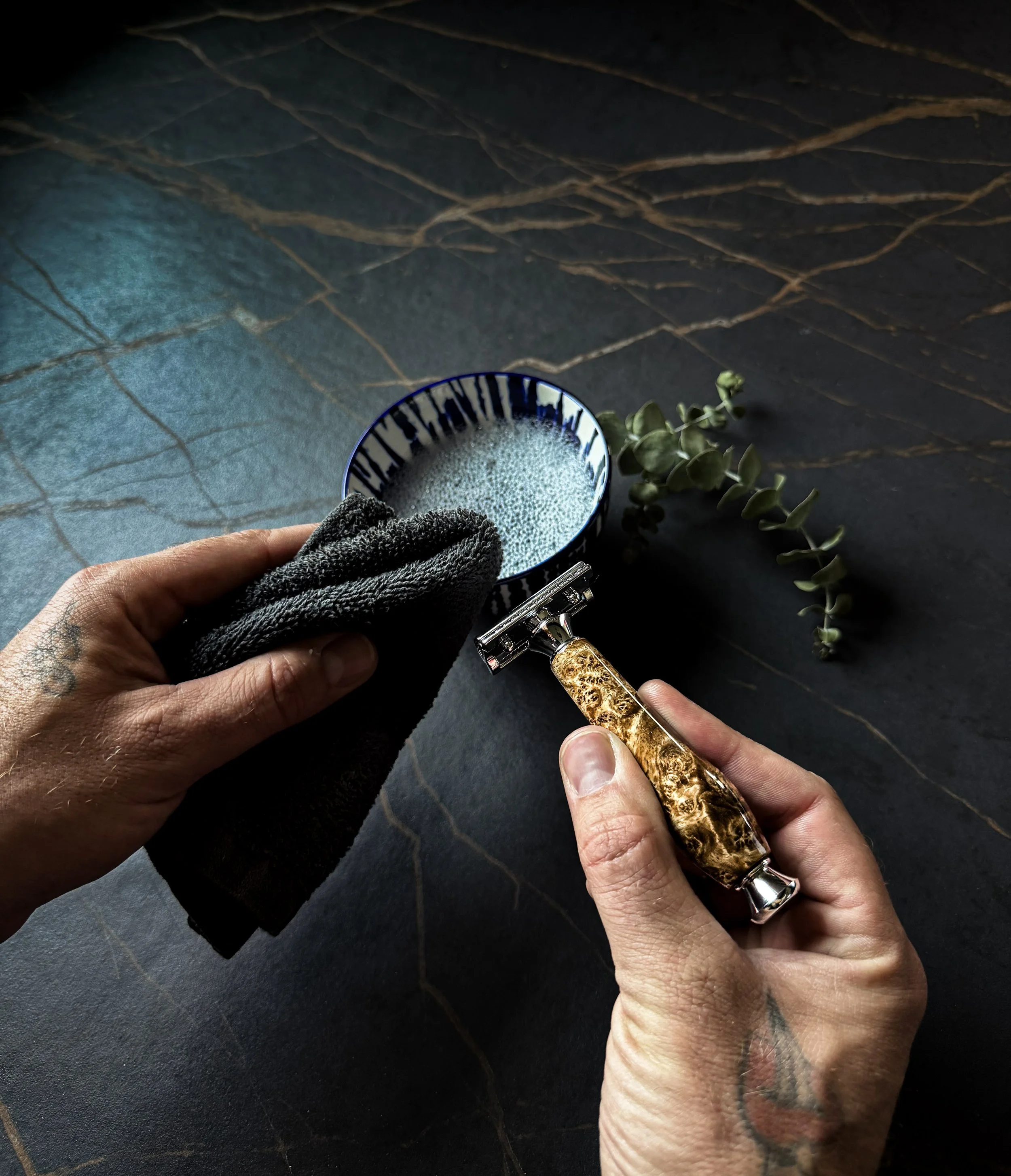 A person's hand holding a shaving razor with a decorative burl handle, next to a small bouquet of green leaves, with a black textured surface and a blue bowl with white bubbles in the background. blackburl works