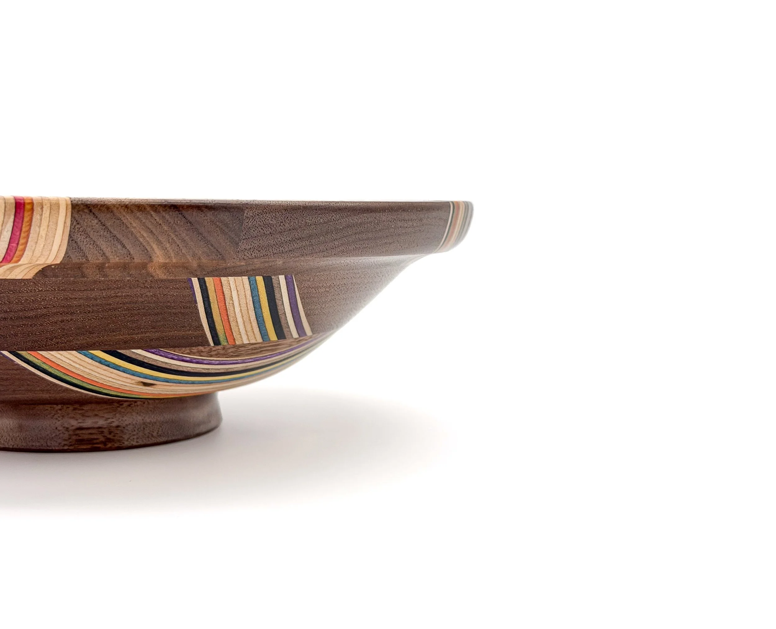 Close-up side view of a wooden bowl with colourful recycled skateboard striped patterns, placed on a white surface.