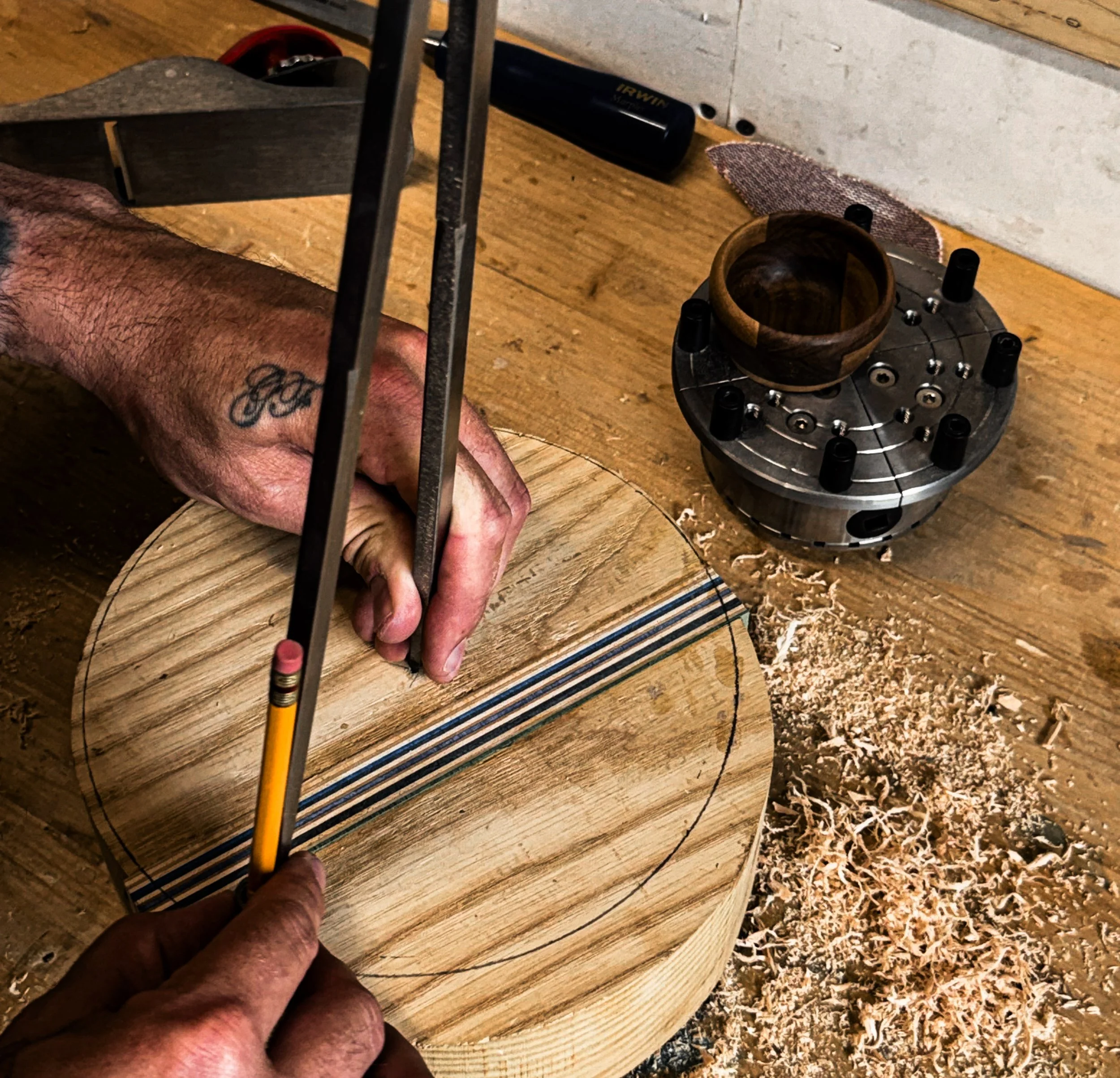 Close-up of a man's hand holding a carving tool, working on a piece of wood with a circular template and wood shavings on the surface.