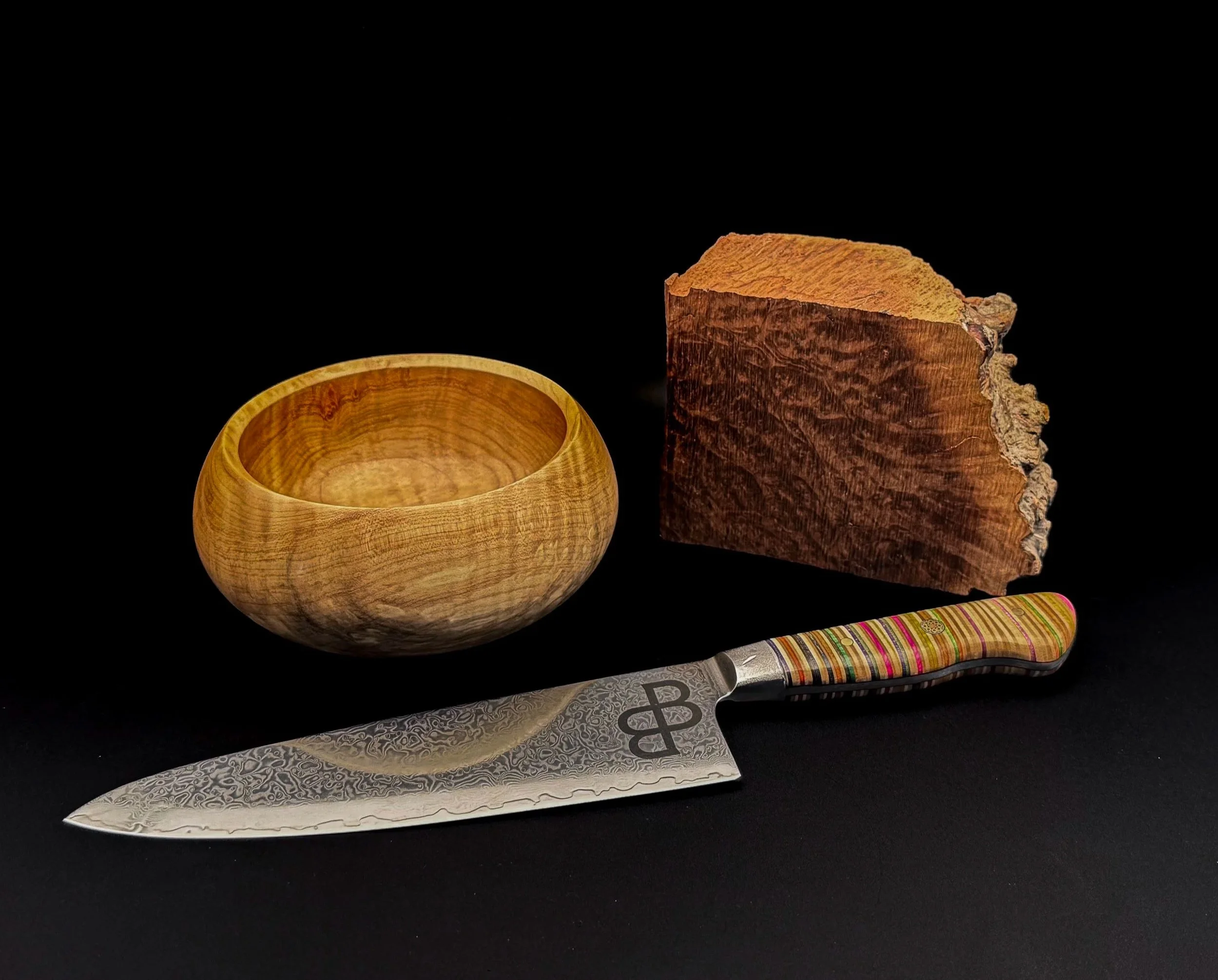 A handcrafted Damascus chef knife with a recycled skateboard handle displayed beside a hand-turned wooden bowl and a block of curly maple burl on a black background.