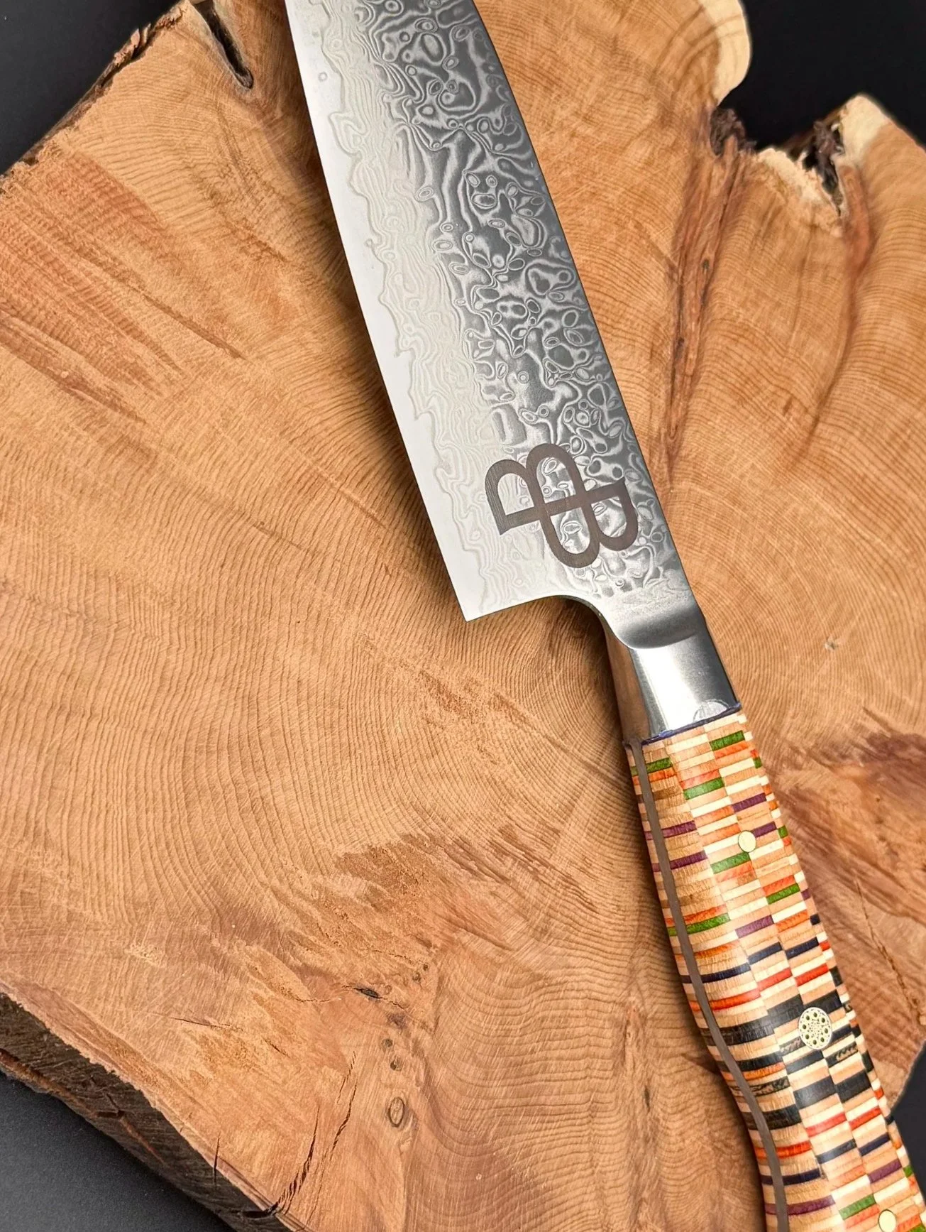A close-up photo of a handmade kitchen knife resting on a large wooden chopping block. The knife has a patterned steel blade with a logo and a multicolored recycled skateboard handle.