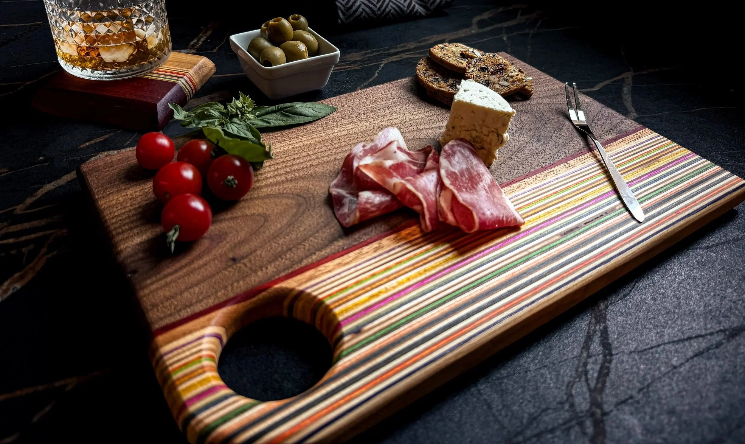 A handcrafted walnut serving board with a recycled skateboard inlay styled with cured meats, blue cheese, cherry tomatoes, olives, basil, and artisan crackers on a dark stone surface.