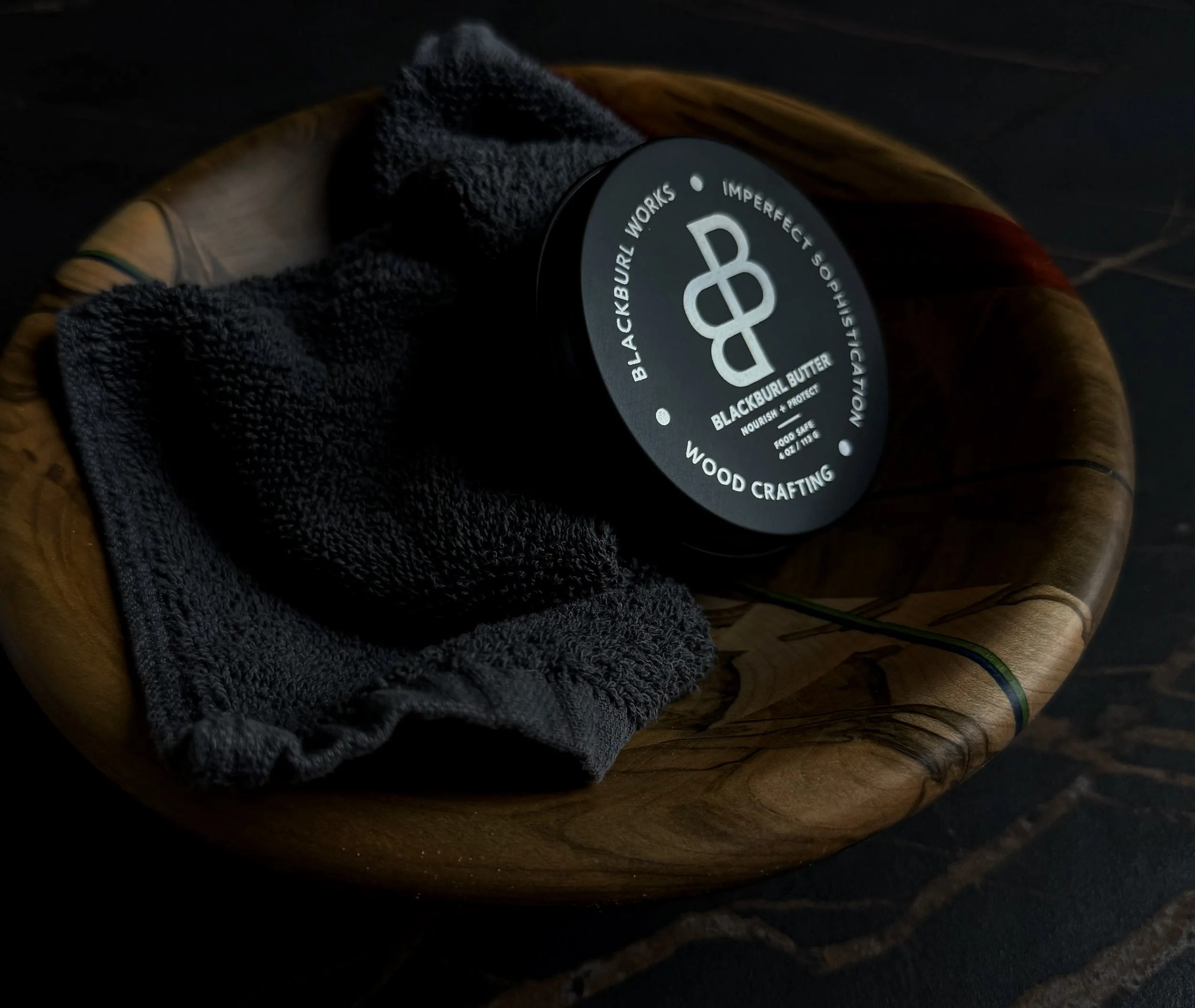 A tin of Blackburl Butter wood conditioner resting in a handcrafted wooden bowl with a dark polishing cloth on a black stone surface.