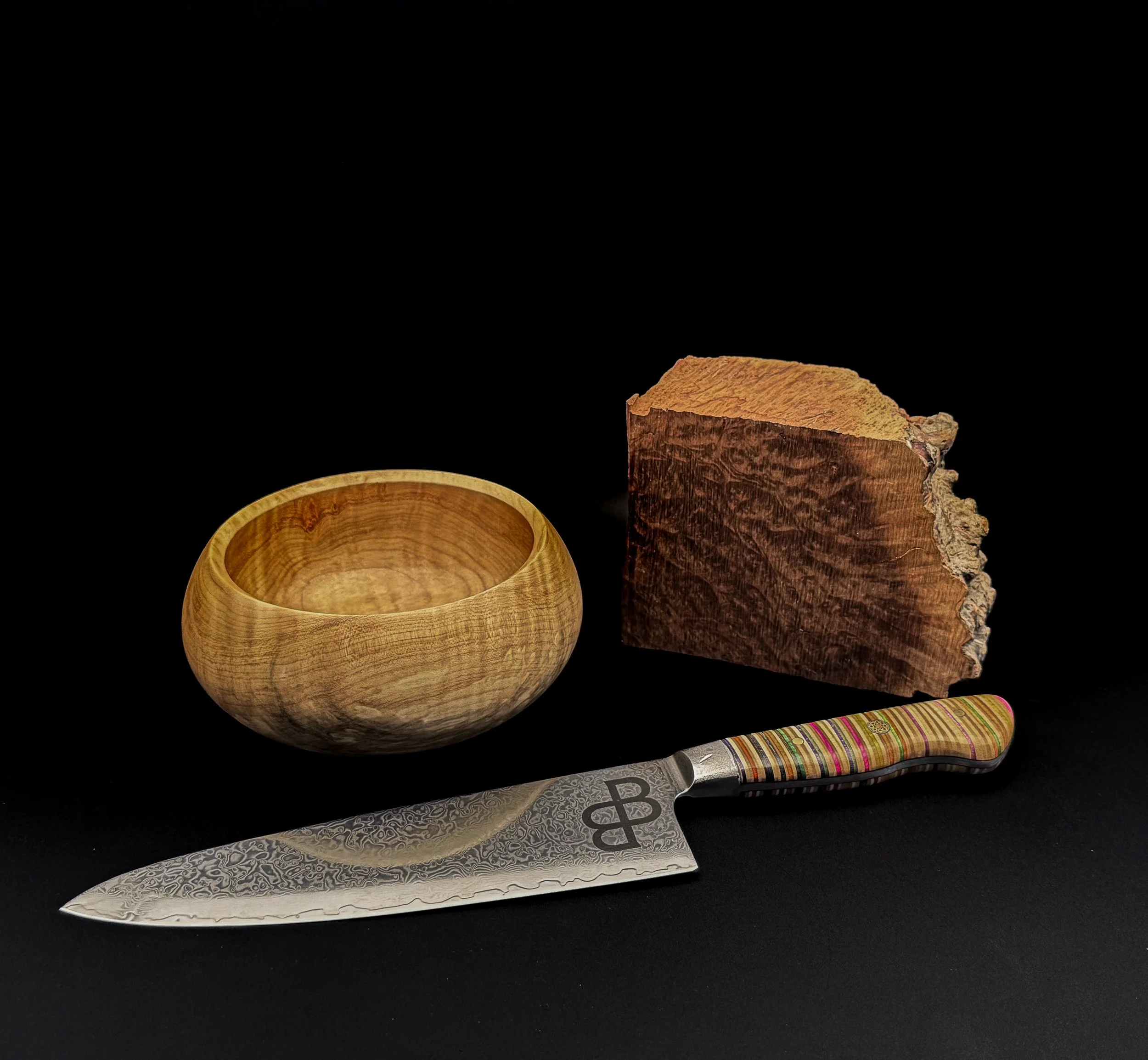 A wooden bowl, a piece of wood, a knife with a colorful striped handle, and a patterned metal knife on a black background.