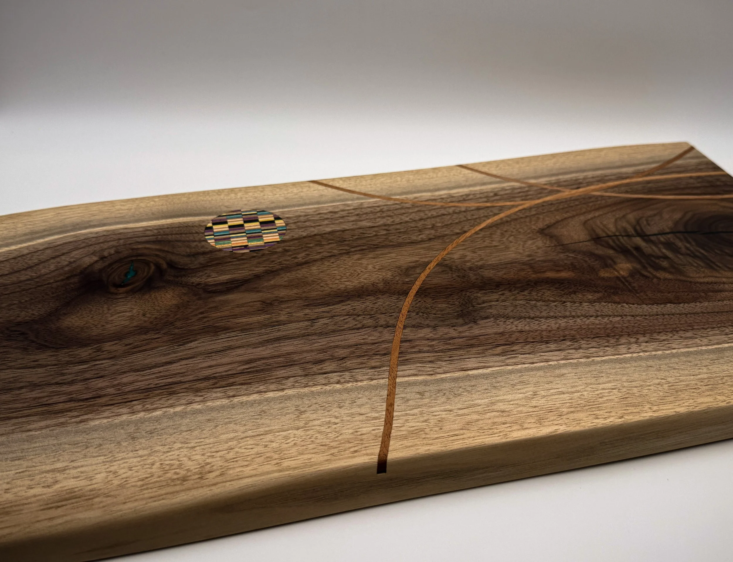 A live egde wooden serving board with a circular multicolored recycled skateboard mosaic inlay.  Wooden bending mahogany inlay.