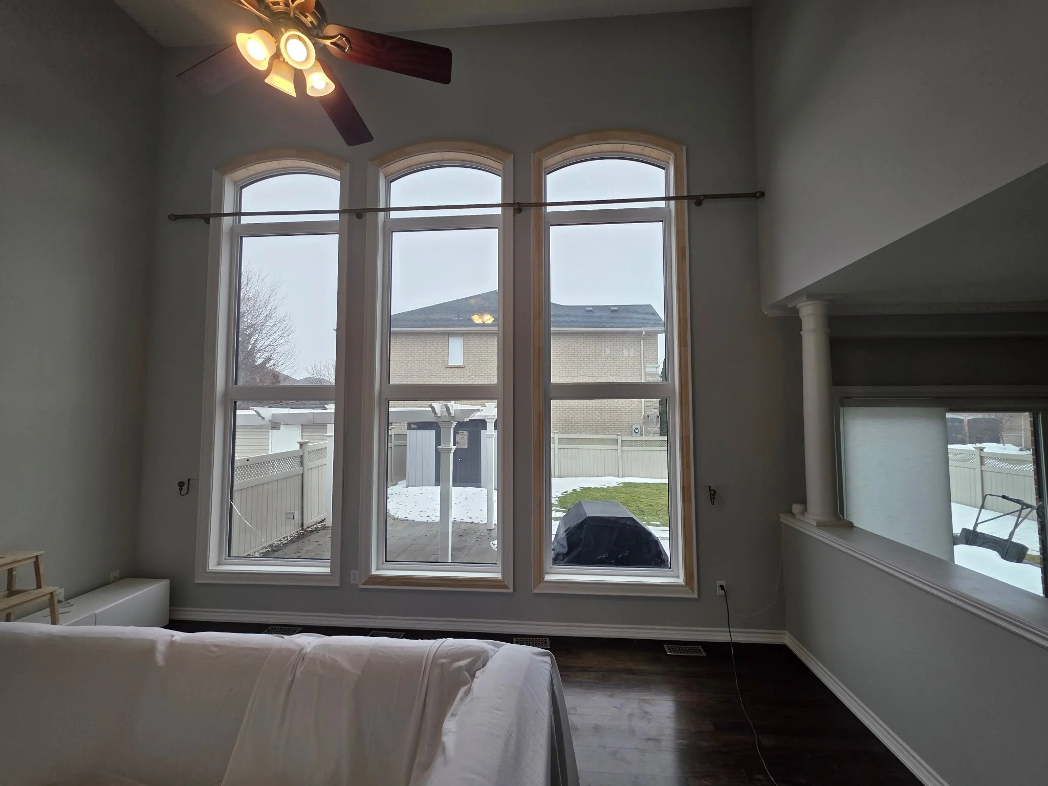 Living room with three tall arched windows showing an outdoor backyard with a small shed and snow-covered ground, a ceiling fan with lights, and part of a white sofa.
