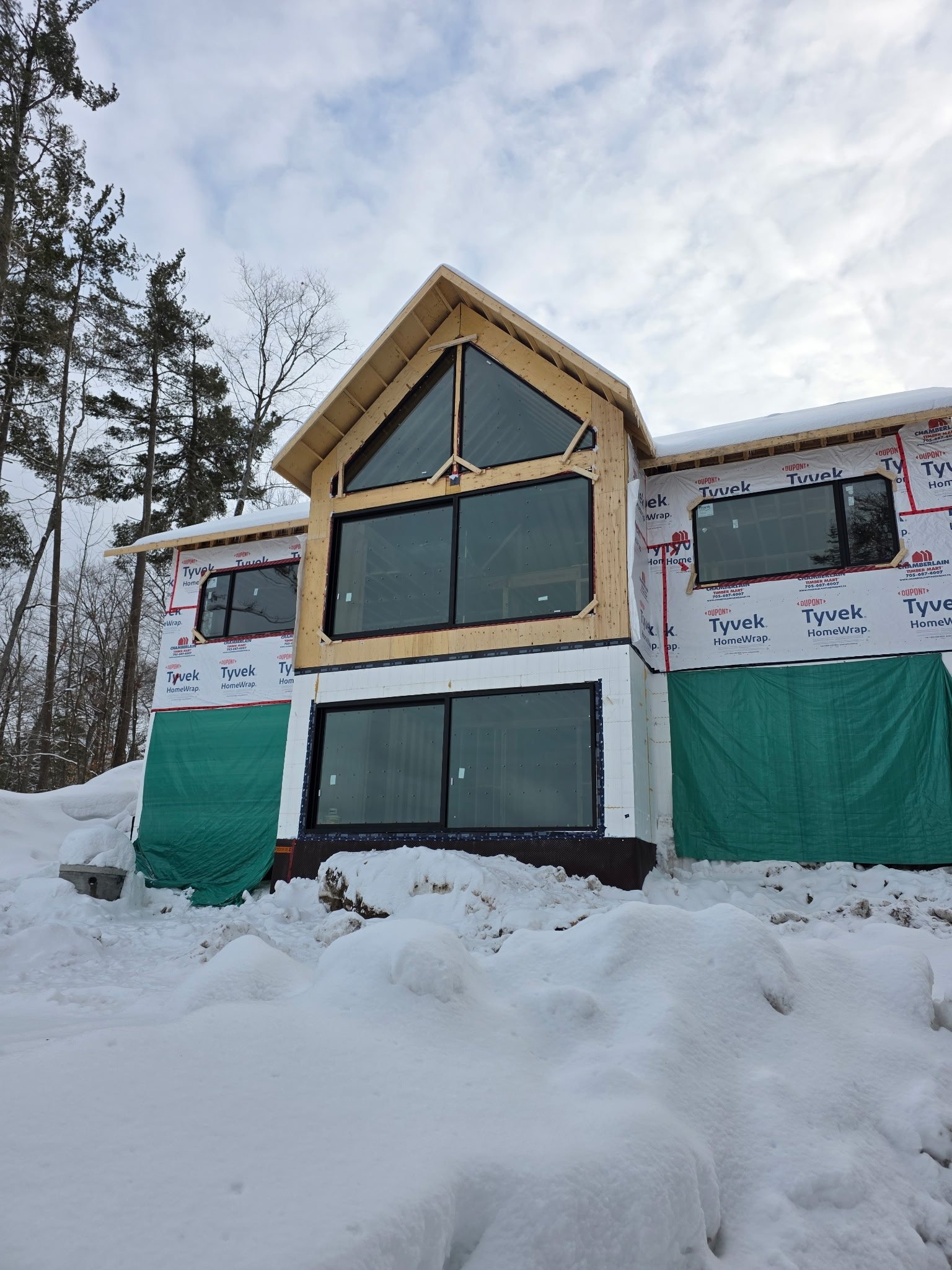 A house under construction with snow on the ground and trees in the background, featuring large windows and exterior insulation wrap.
