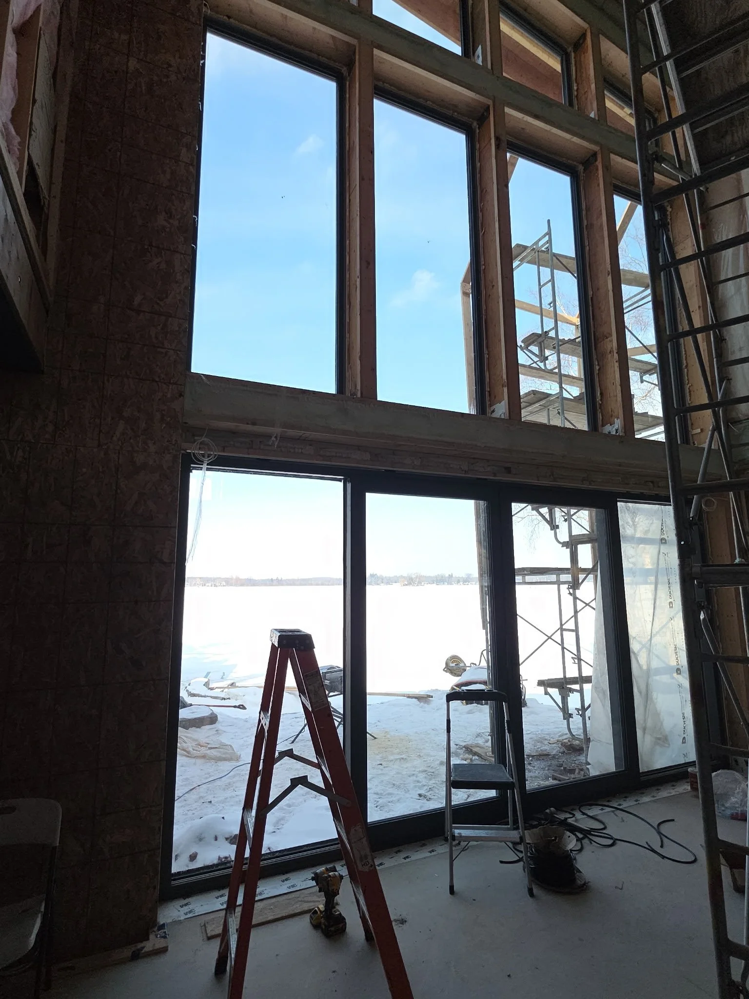 Interior of a building under construction with large window frames, a red step ladder, a black chair, construction tools, and scaffolding outside visible through the window.