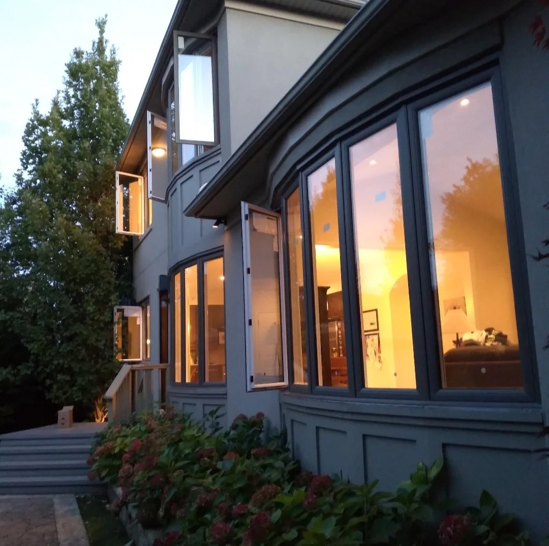 A multi-story house with large, open windows illuminated from inside during sunset, surrounded by trees and bushes.