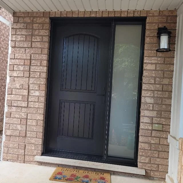 Black front door with a sidelight window on the right, brick exterior walls, a wall-mounted outdoor lantern to the right of the door, and a welcome mat in front that says "Gnome Grown" with gnome and mushroom illustrations.