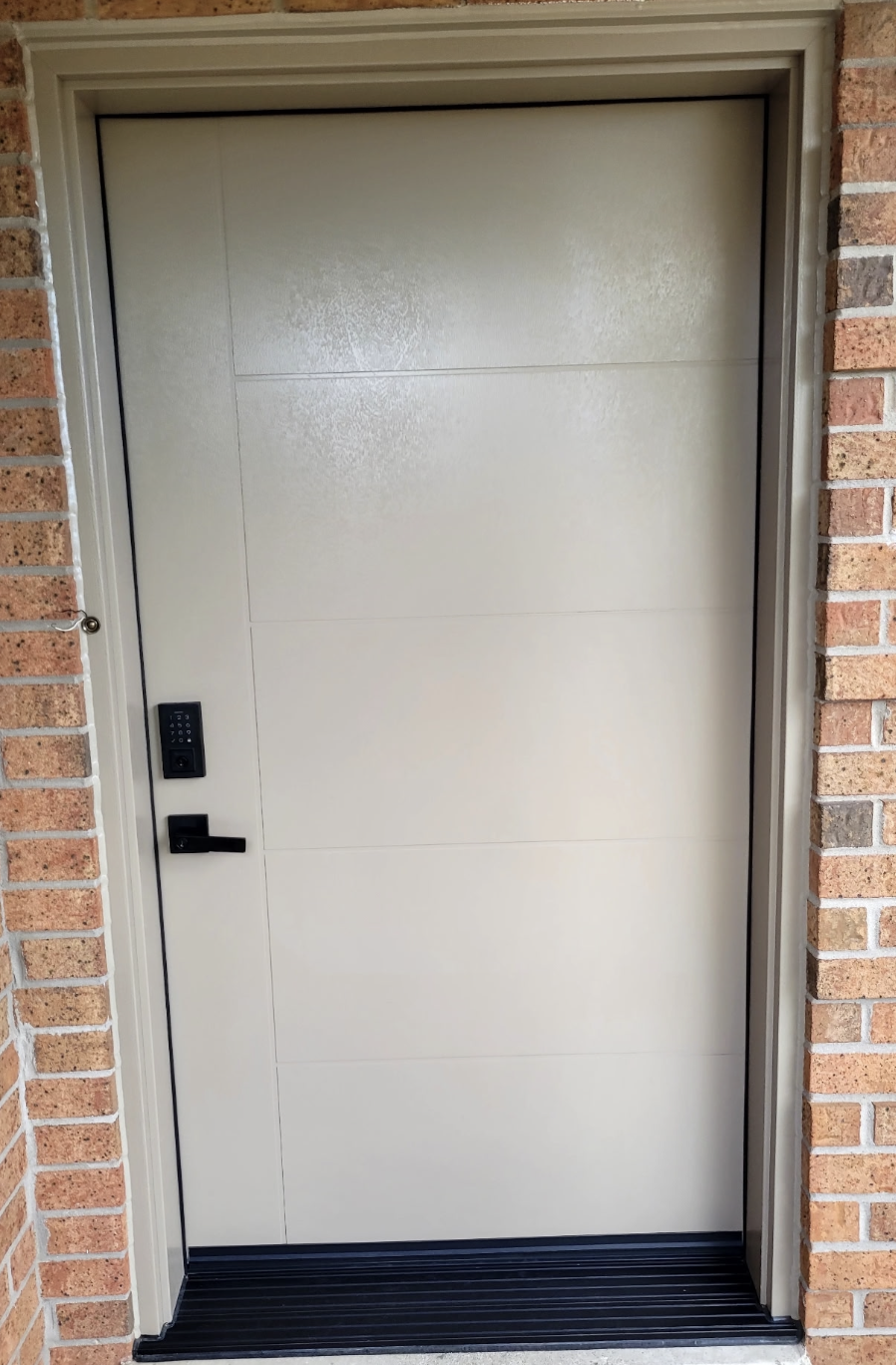 A modern, light-colored garage door with a digital keypad lock and a black handle set in a brick wall.