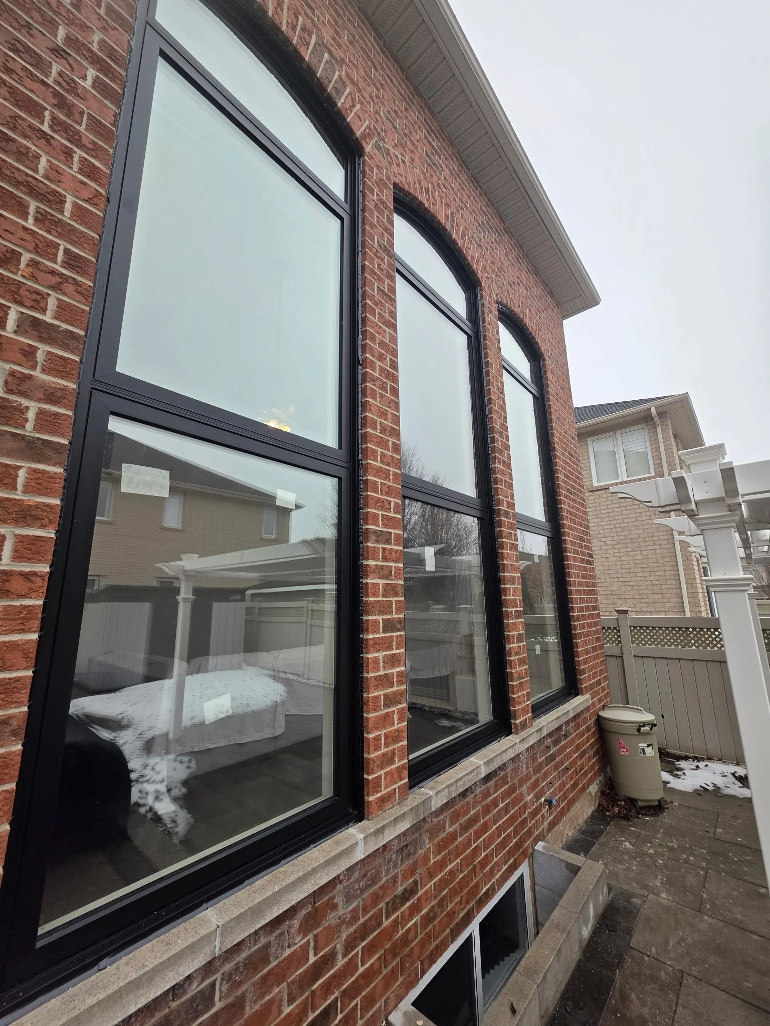 The image shows the exterior of a house with red brick walls and large black-framed windows. The house has a white overhang roof, and the ground is concrete with some scattered leaves and snow. In the background, parts of neighboring houses are visible.