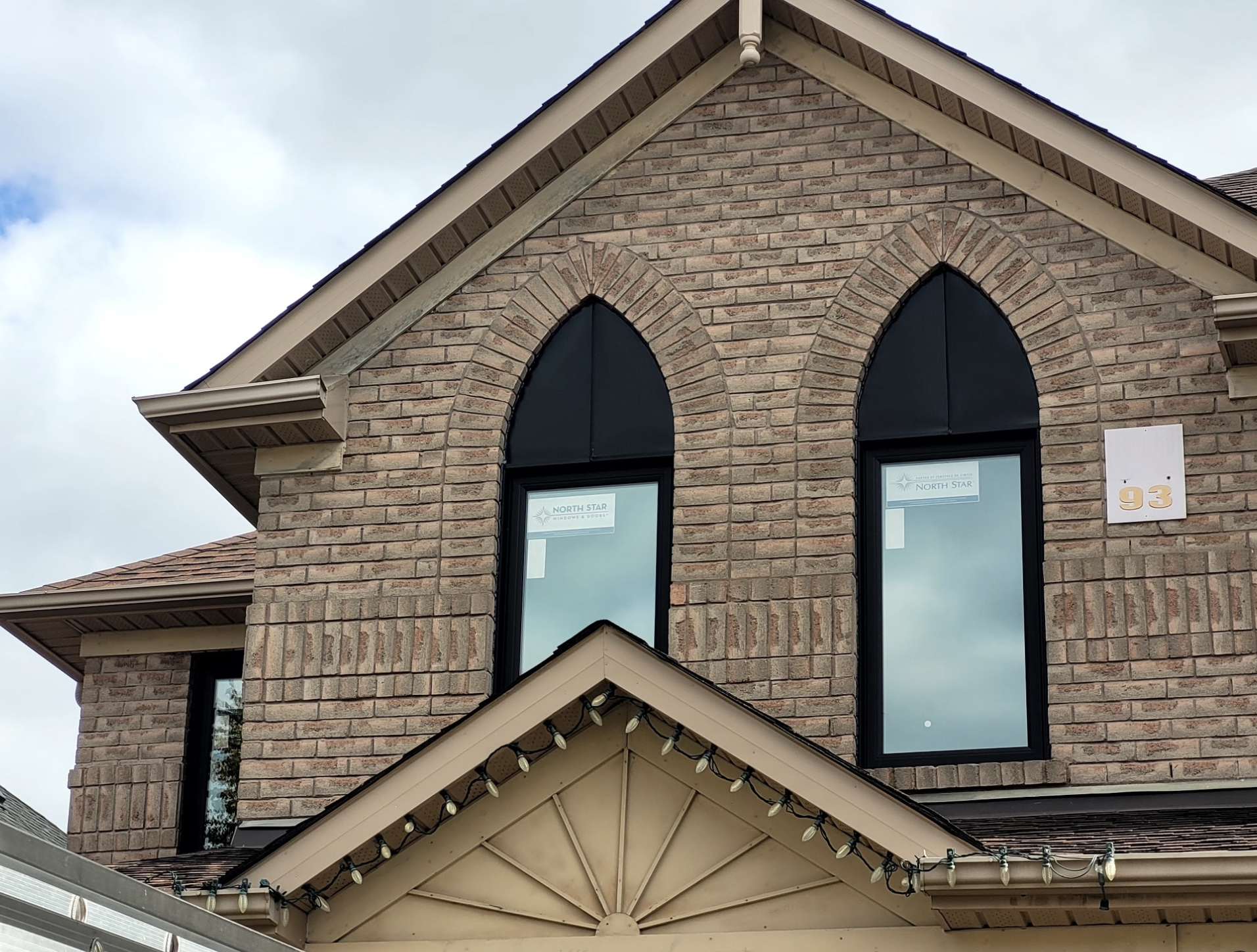Close-up of a brick house's upper story with two pointed arch windows with black frames, a beige trim, and a house number 93 on a white plaque, along with Christmas string lights hanging from the roof.