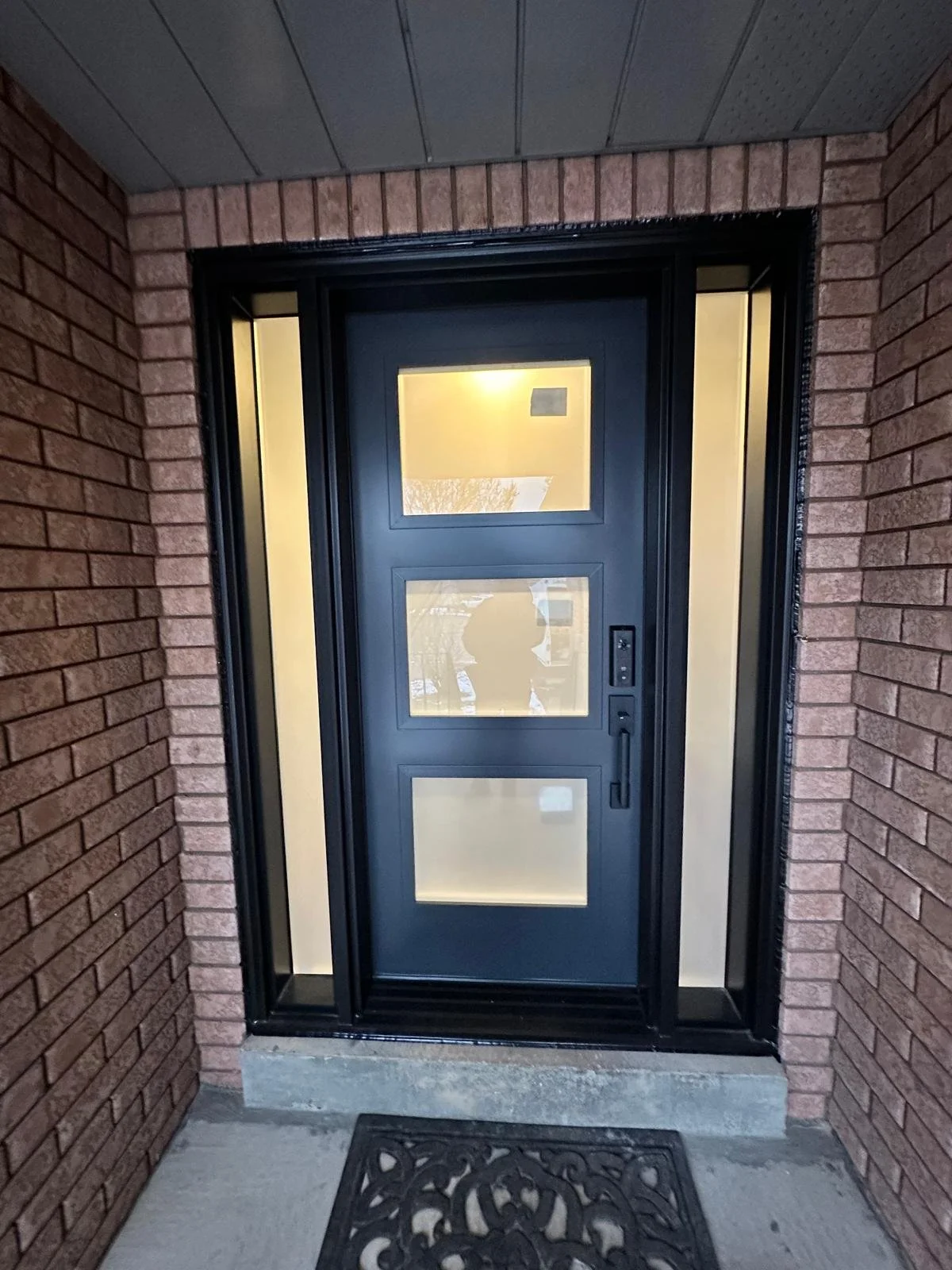 Black front door with three rectangular glass windows, flanked by sidelight windows, set in a brick house entryway with a black decorative doormat.
