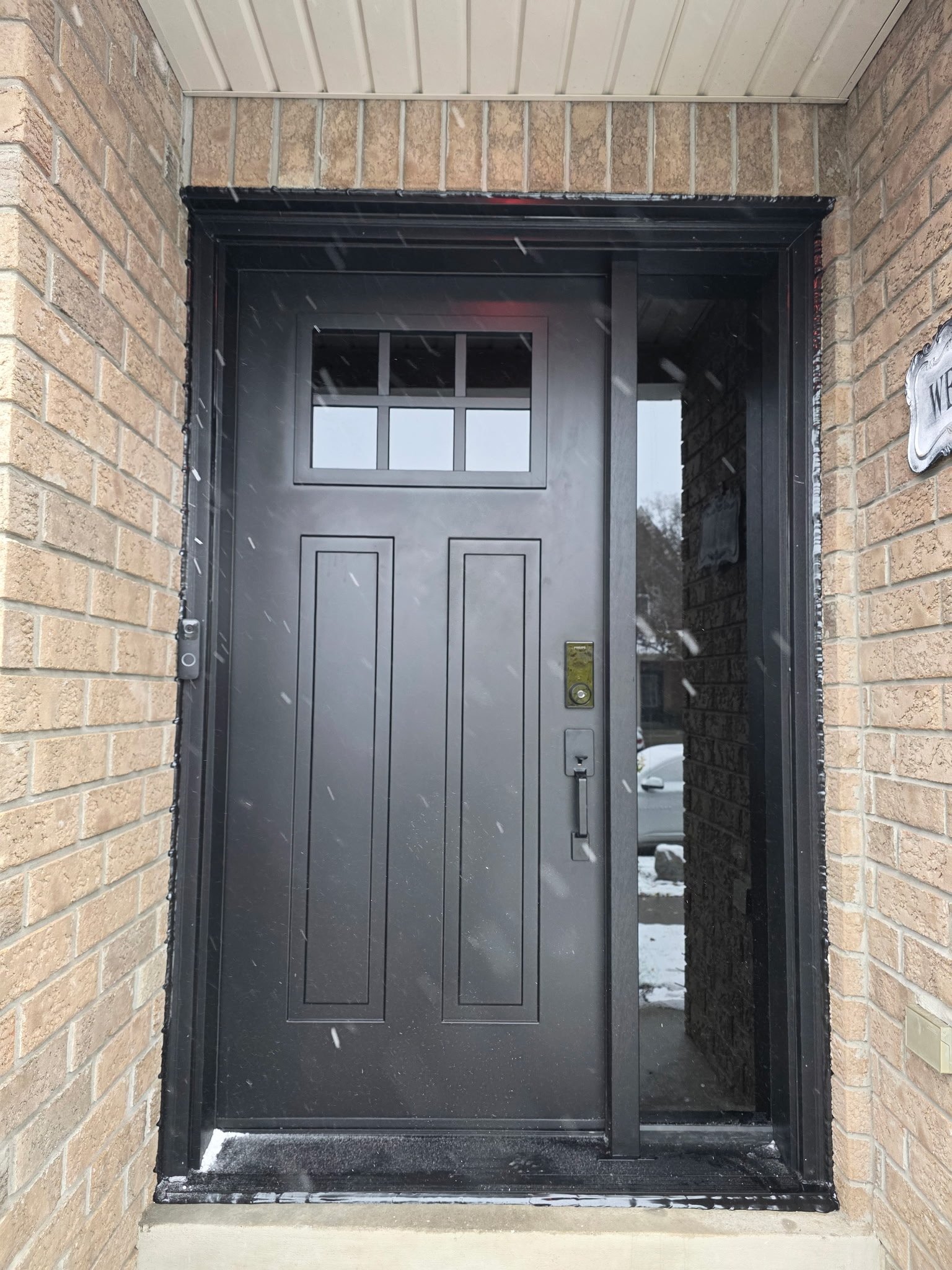 Black front door with a window panel at the top, surrounded by brick walls and an opaque glass side panel, during snowy weather.