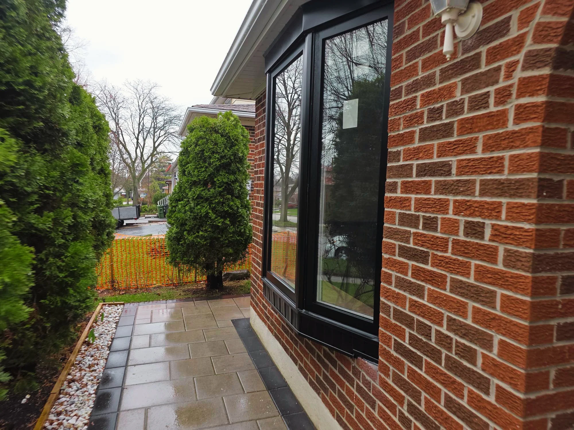 View of a house's brick exterior wall with a window, an outdoor light fixture, a paved patio, and green shrubs in a suburban neighborhood with leafless trees in the background.