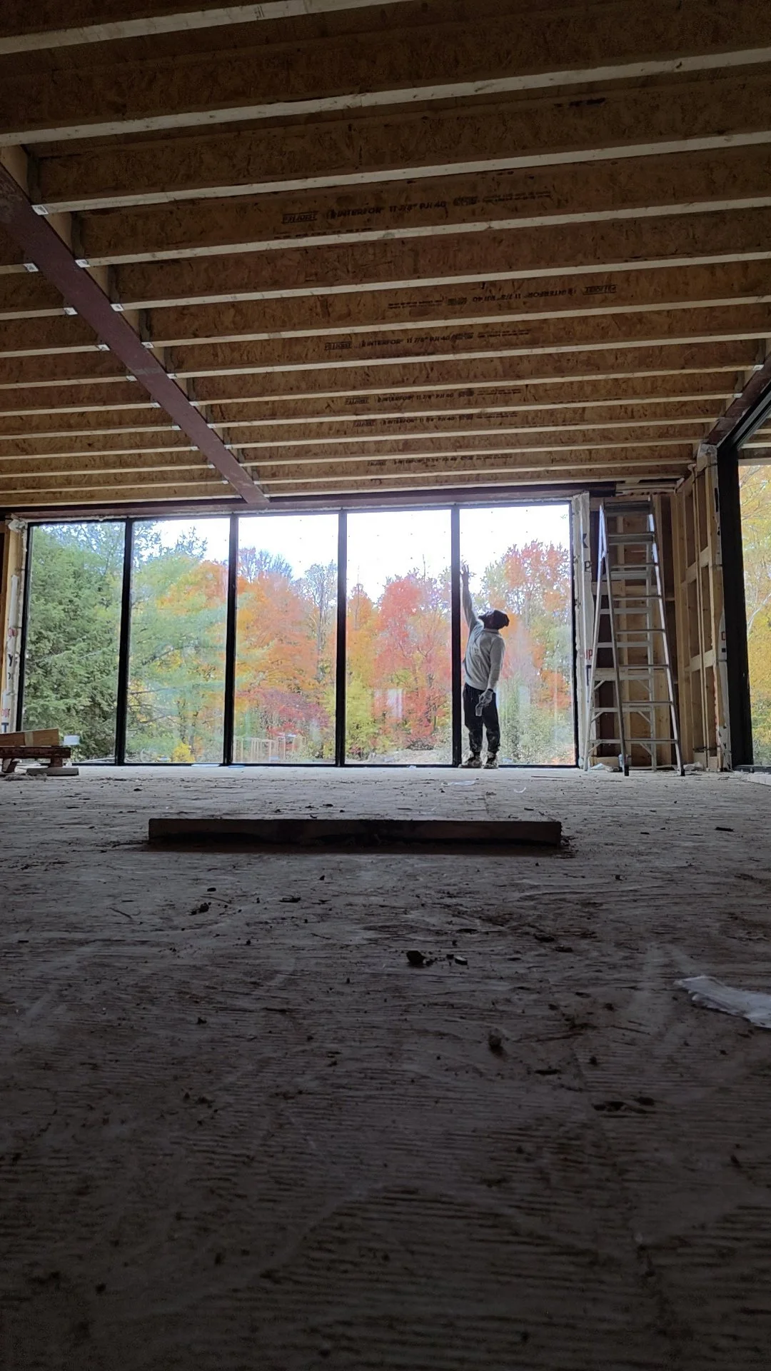 Interior view of a building under construction, with a worker standing near large window frames and looking outside at autumn trees.