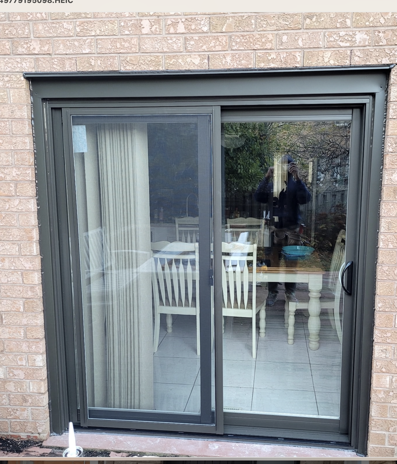 A sliding glass door with a black frame, leading into a dining room with white chairs around a wooden table, seen through the glass are reflections of trees and a person taking the photo.