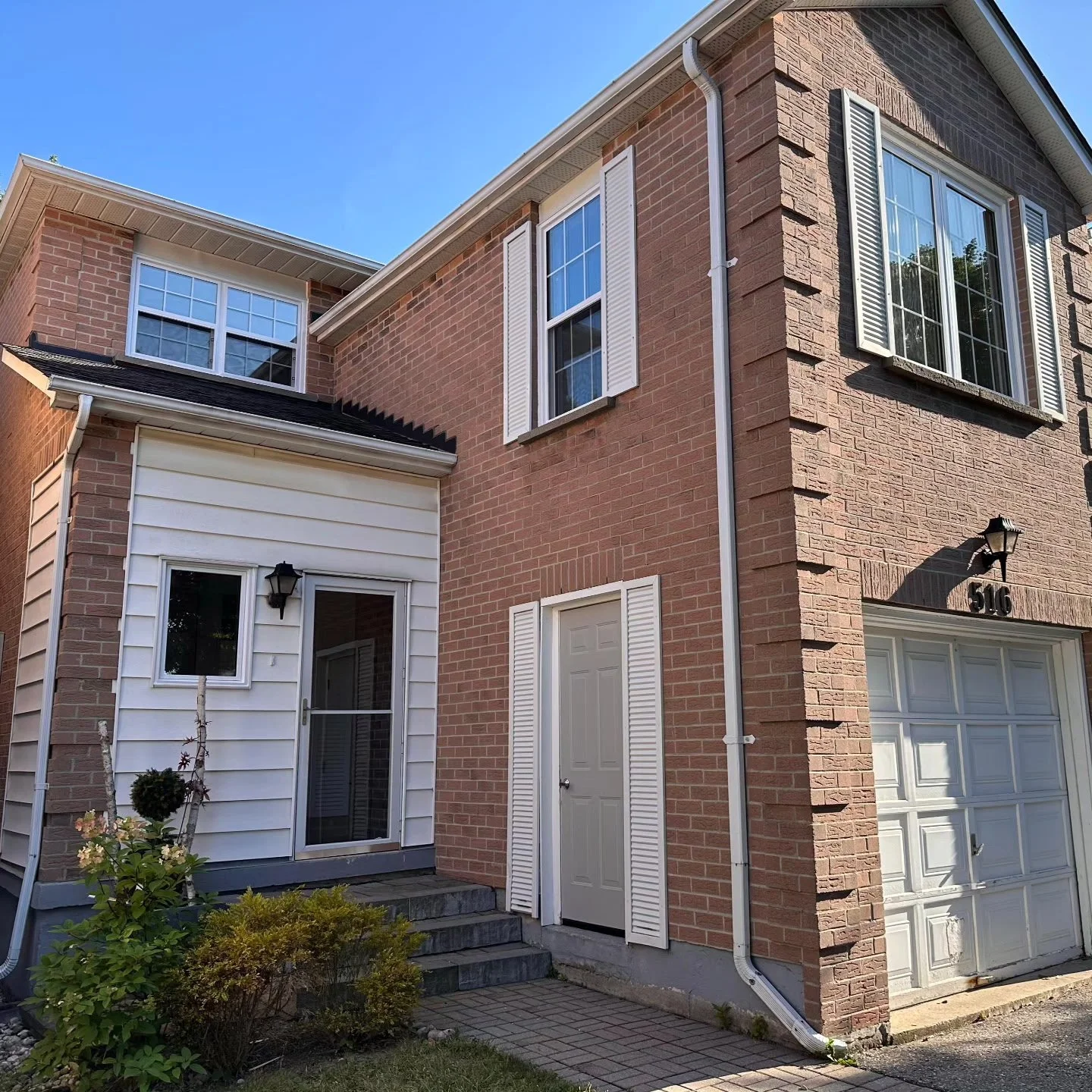 Front facade of a two-story brick house with white trim and a garage door, featuring windows with white shutters and a small porch with steps leading to the entry door, under a clear blue sky.