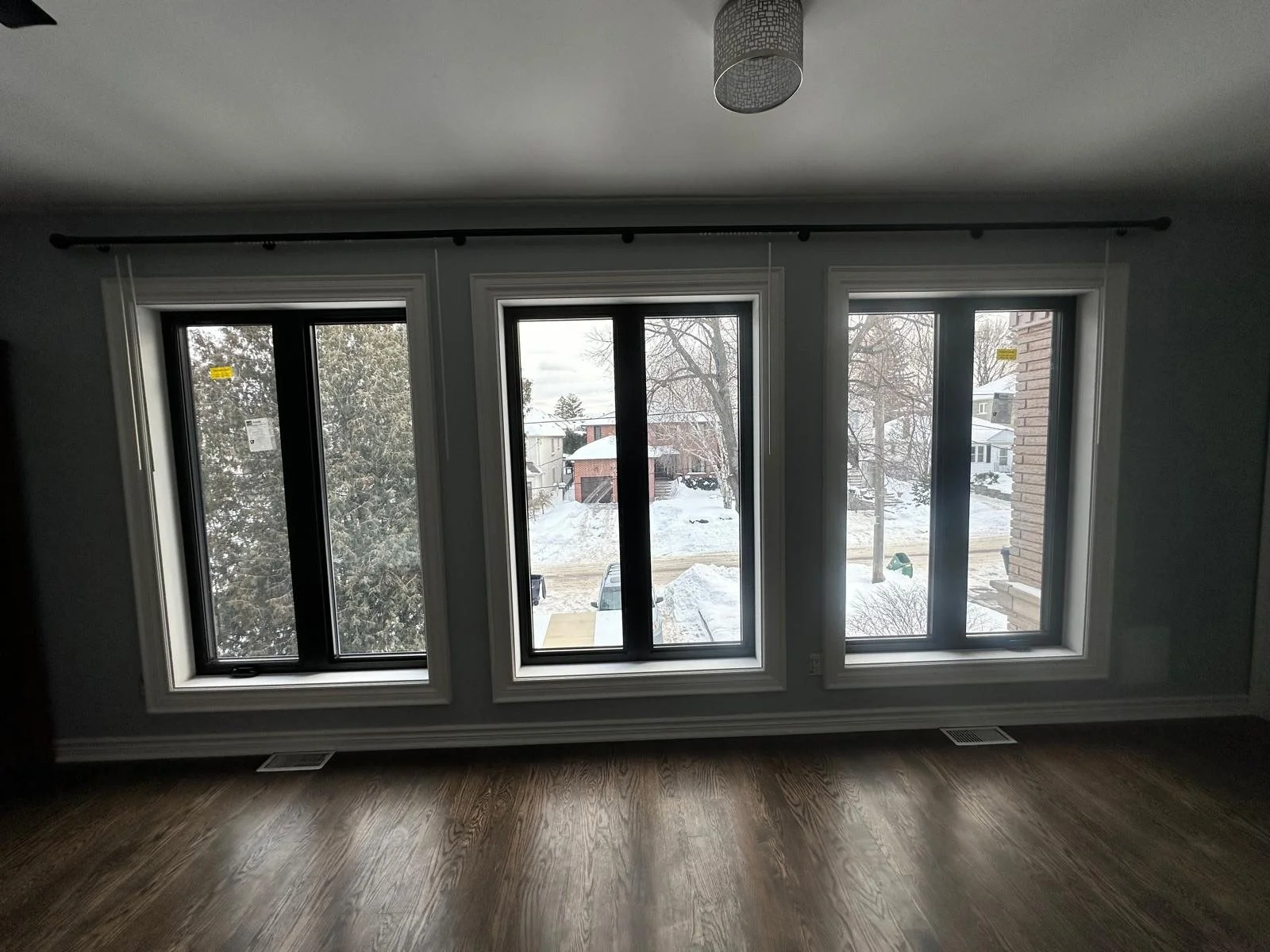 A room with three large windows overlooking a snowy landscape with trees, houses, and parked cars.