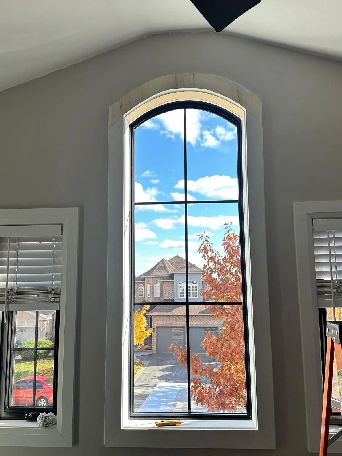 Interior view of a large, arched window with black grid panes, showing a neighborhood with a house, driveway, and trees with autumn leaves outside on a sunny day