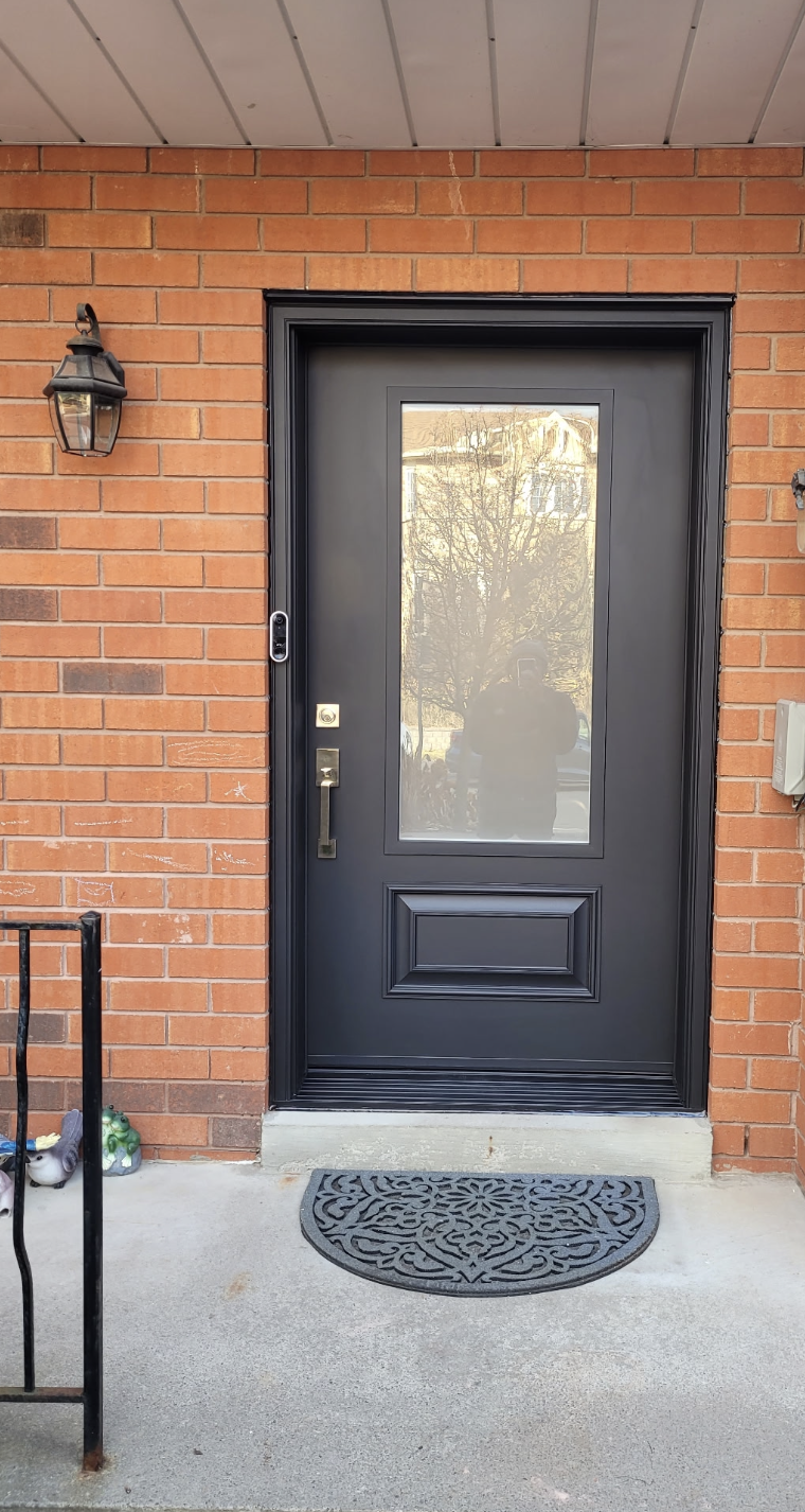 Front door of a house with a red brick exterior, black door with a glass window, a wall-mounted lantern light, a black metal railing, and a decorative black doormat.