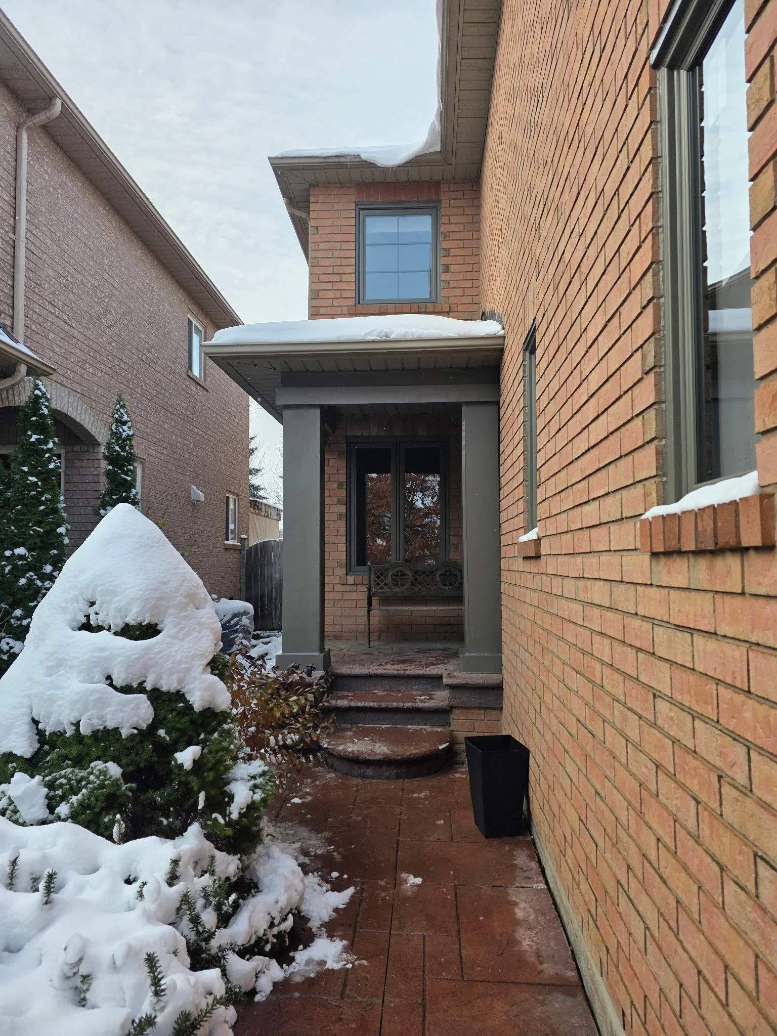 A backyard entrance with snow-covered plants, brick pathway, and a brick house wall with large windows and snow on the roof and ground.