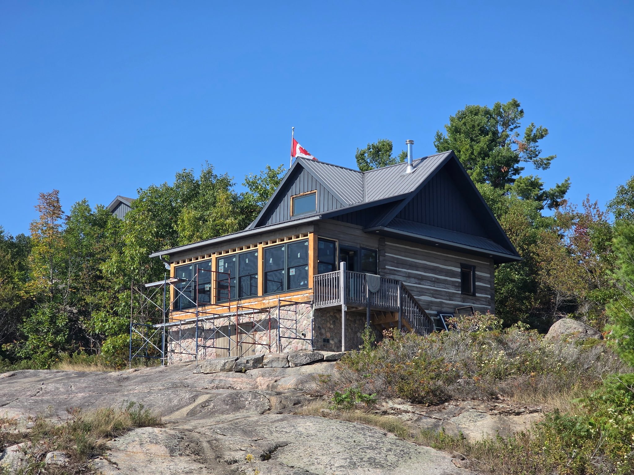 A house on a rocky hill with trees behind it, a Canadian flag on the roof, large windows on the porch, and a clear blue sky.
