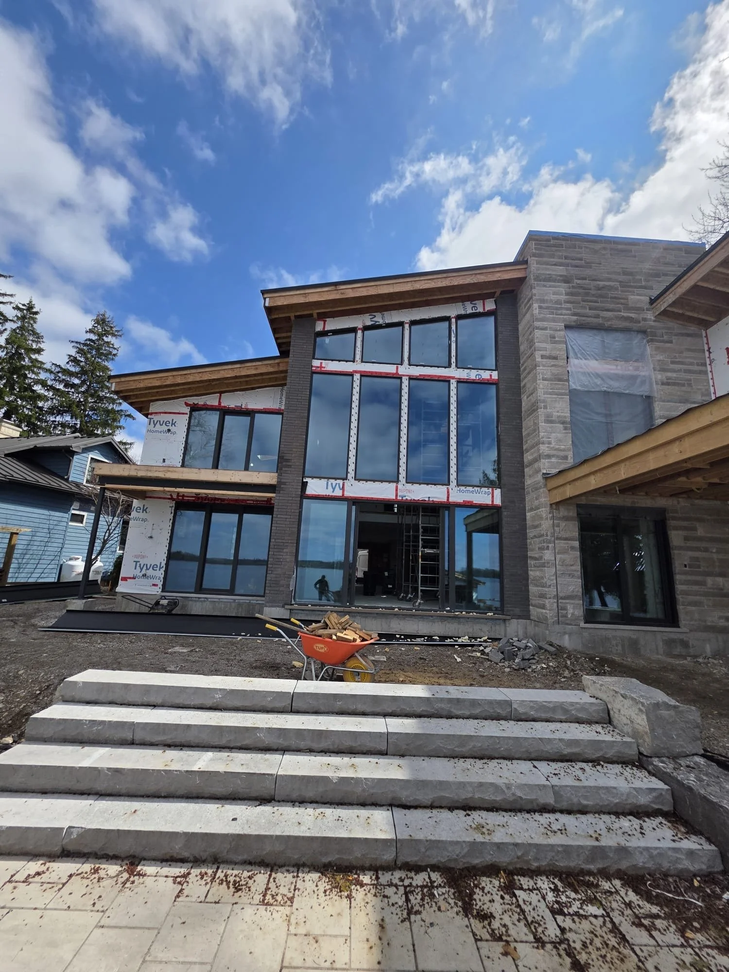 A multi-story house under construction with large glass windows and a stone exterior, set against a partly cloudy blue sky, with stairs and construction materials in the foreground.