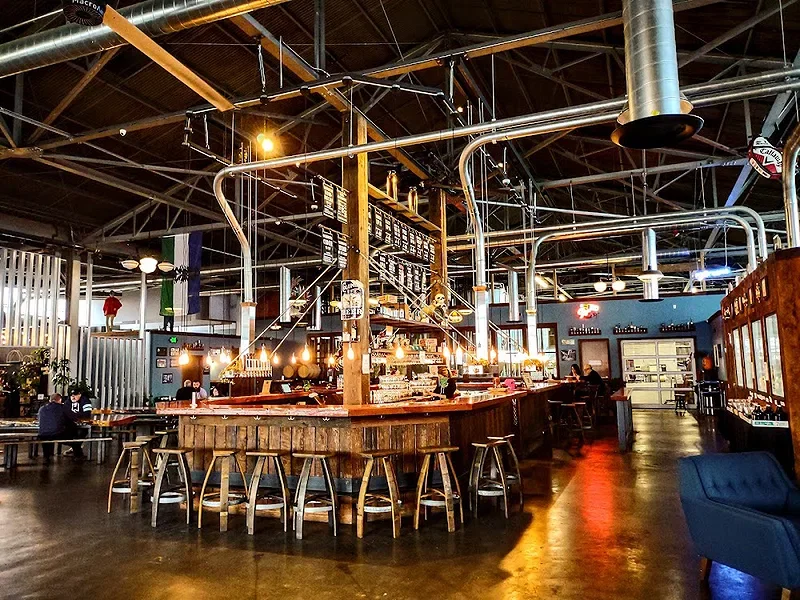 Interior of a modern industrial-style bar with a large curved wooden bar counter, high stools, and exposed pipes and ductwork on the ceiling. There are some patrons sitting at the bar, and other tables with people in the background. The lighting is warm and ambient.
