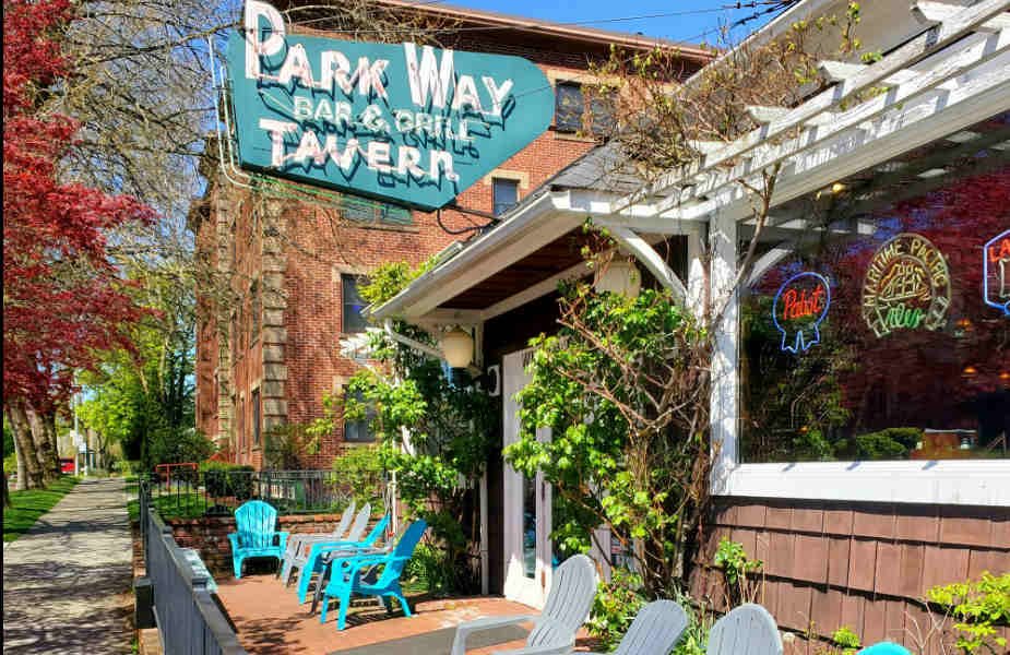 Exterior of Park Way Tavern with outdoor seating area, blue and white chairs, hanging neon signs, and a large signboard showing the establishment's name.