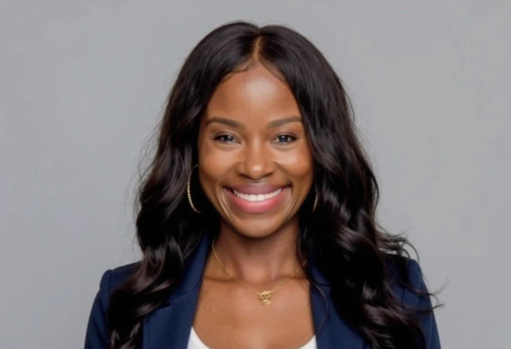 A smiling woman with dark wavy hair, wearing a navy blazer, gold hoop earrings, and a gold necklace, posing against a light gray background.