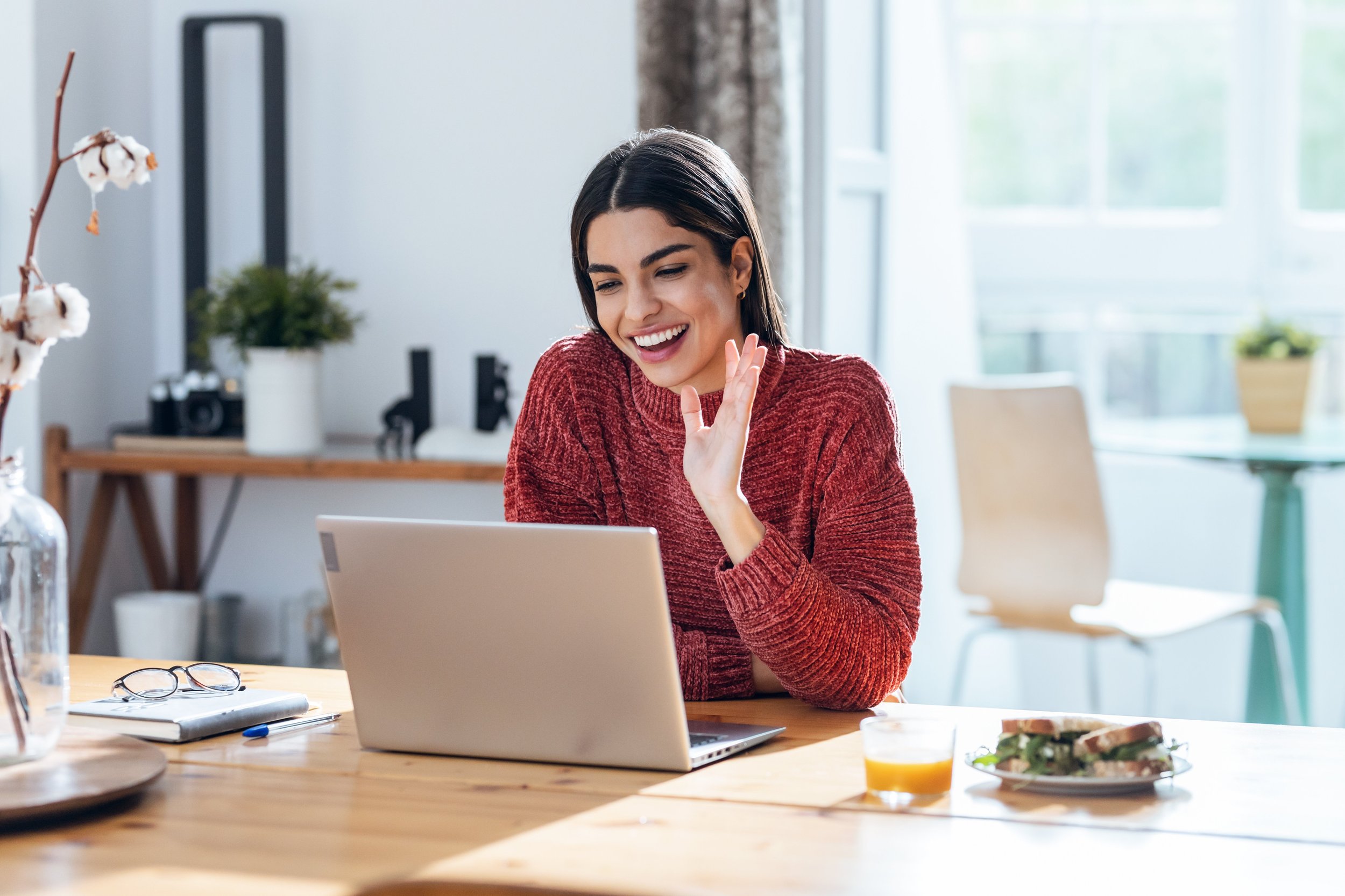 A woman with short dark hair and a red sweater sitting at a wooden table, smiling and waving while looking at her laptop. The table has glasses, a notebook, a pen, a glass of orange juice, and a plate of sandwiches. In the background, there are large windows with curtains and some indoor plants.