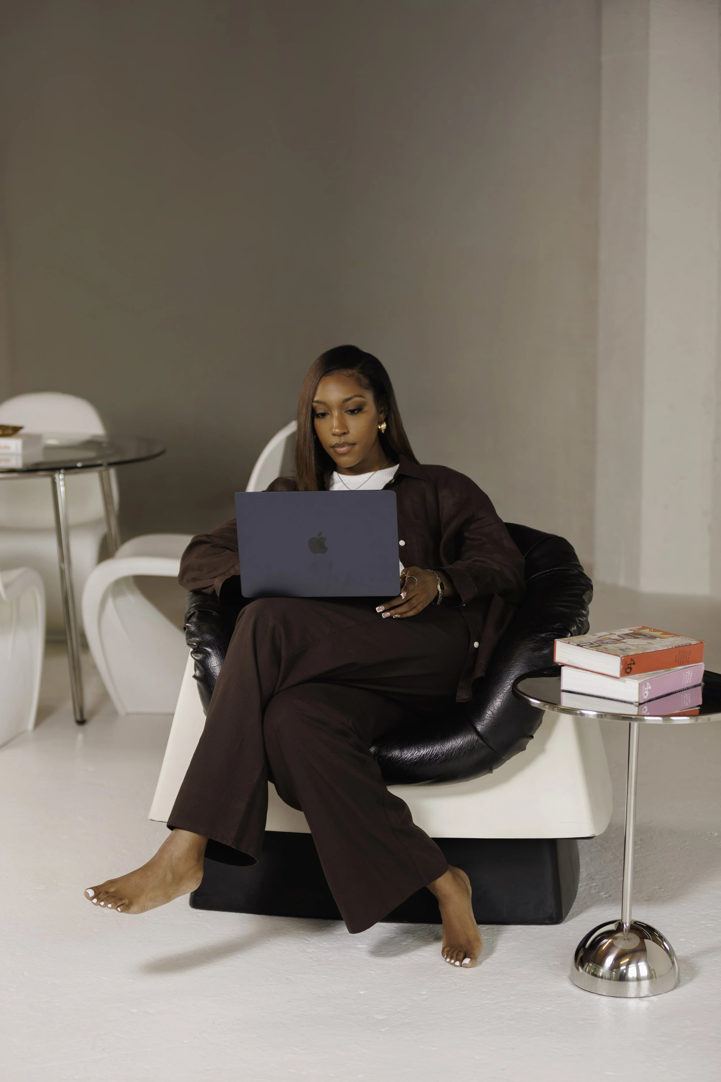 A woman sitting on a black leather chair, barefoot, working on a laptop. She is wearing brown pants and a brown jacket, with a stack of books on a small table next to her. The background includes a white chair, a round glass table, and a beige wall.