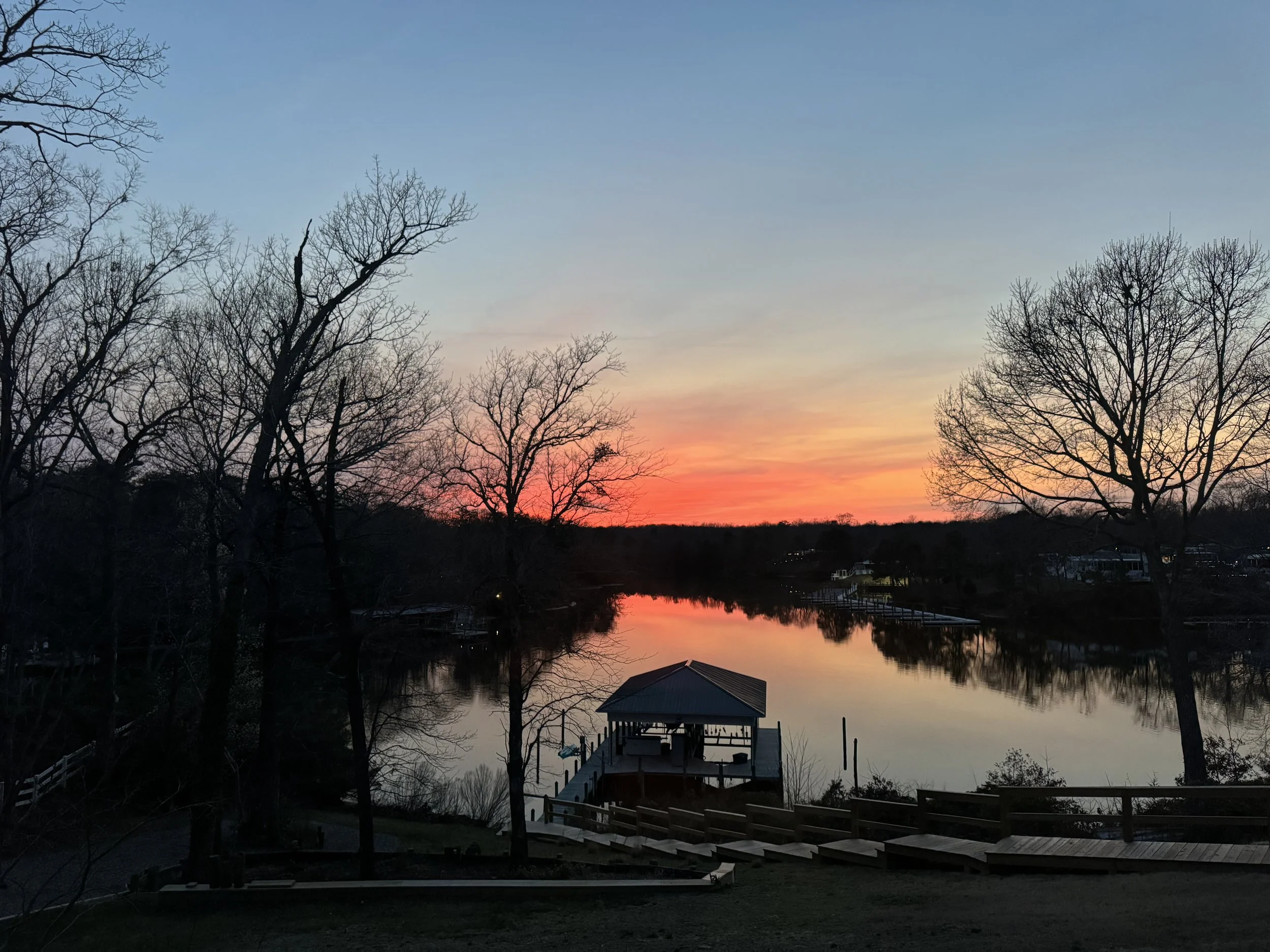 Sunset over a calm river with silhouetted trees on both sides and a small wooden dock with a pavilion in the foreground.