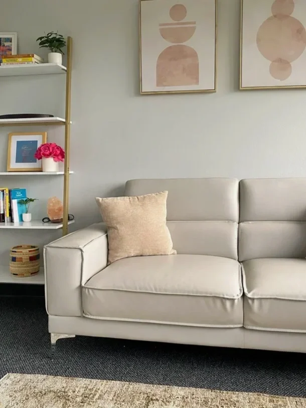 Living room with white leather sofa, beige pillow, black carpet, white wall, abstract art prints, white bookshelf with books, plants, framed picture, pink flowers, salt lamp, woven basket, and a rug.
