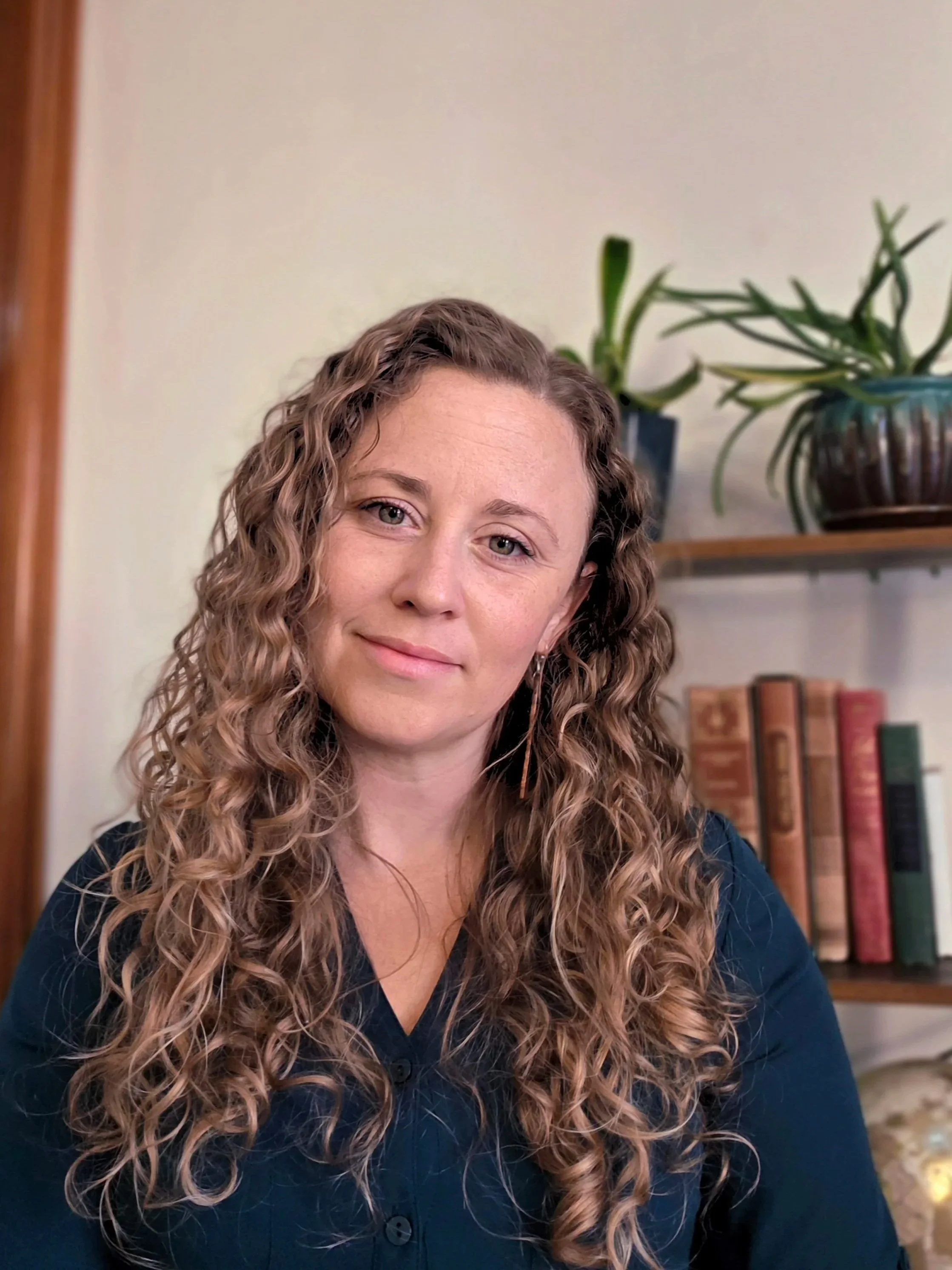 A woman with long, curly hair and light skin, smiling gently, sitting in front of a bookshelf with books and a potted plant.