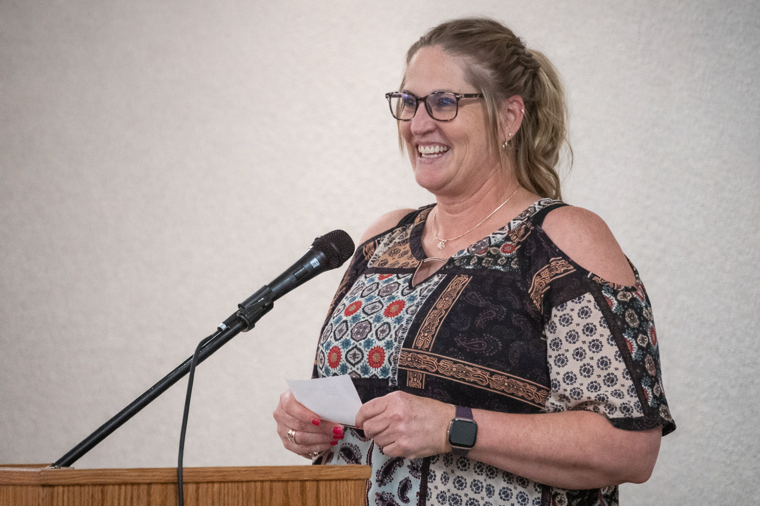A woman with glasses and earrings, smiling, speaking into a microphone while holding a paper.