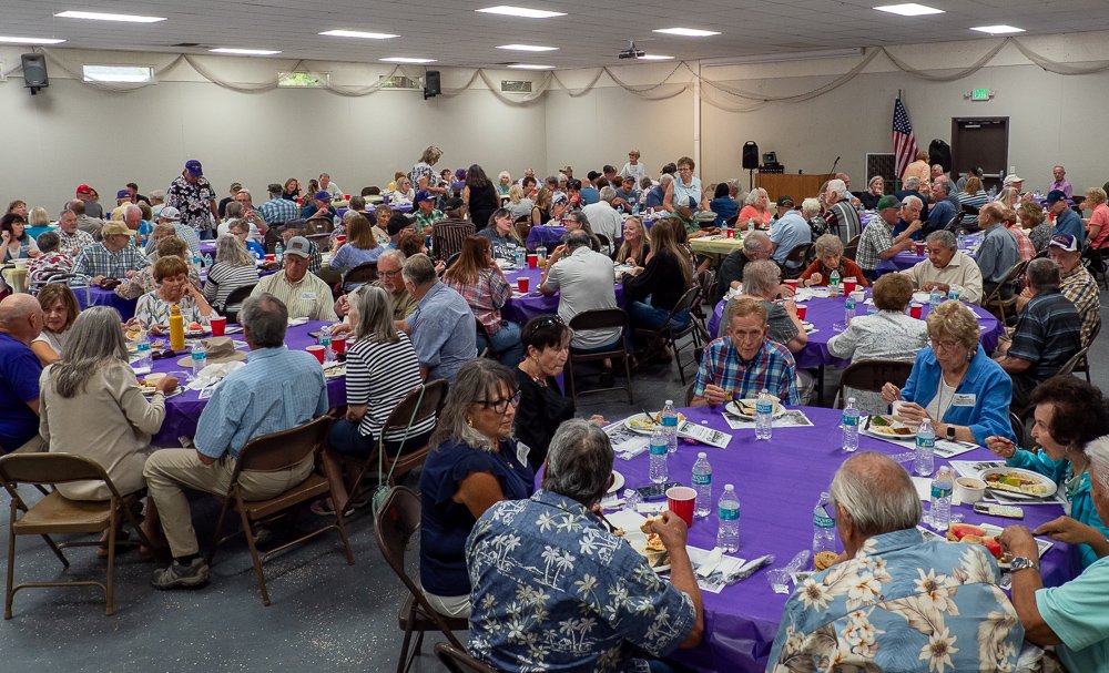 A large gathering of people sitting at tables with purple tablecloths, eating and socializing in a banquet hall. There are many elderly individuals, and the room has an American flag near the front.